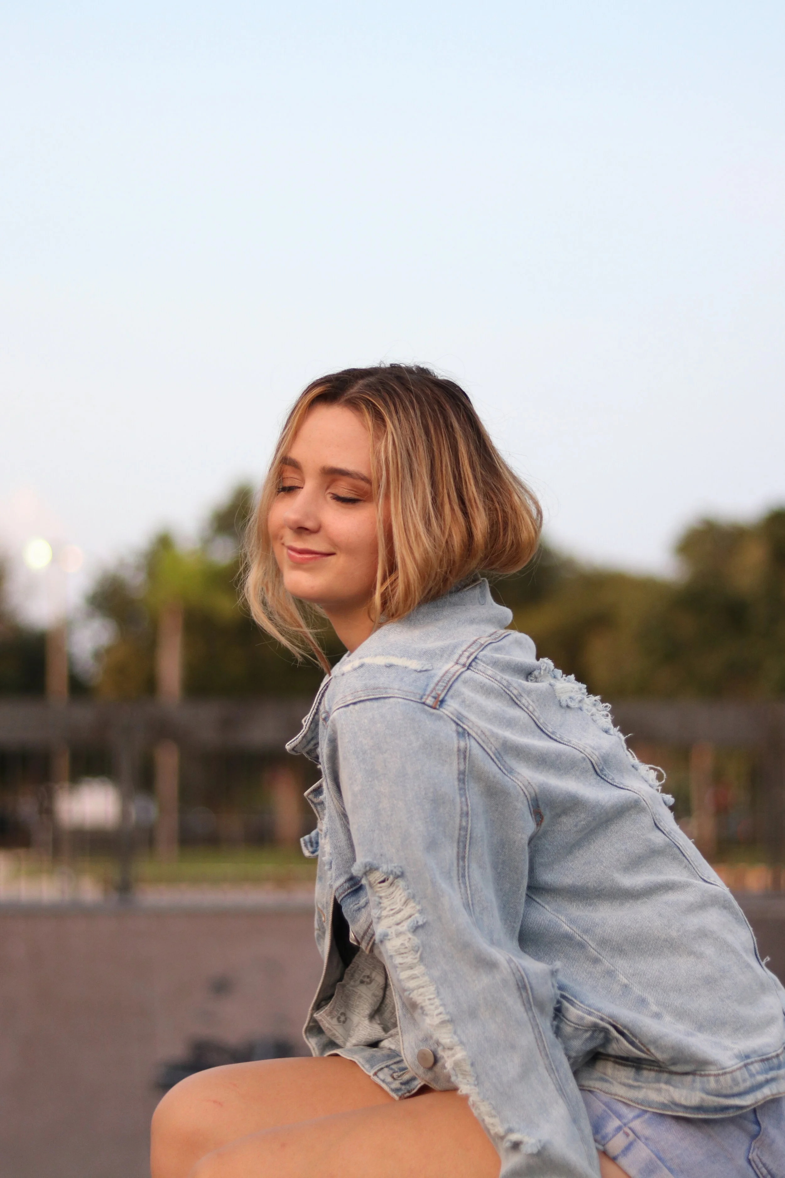 A young woman with shoulder-length blonde hair, wearing a distressed denim jacket, sits outdoors with her eyes closed and a gentle smile, during a clear evening with soft lighting.
