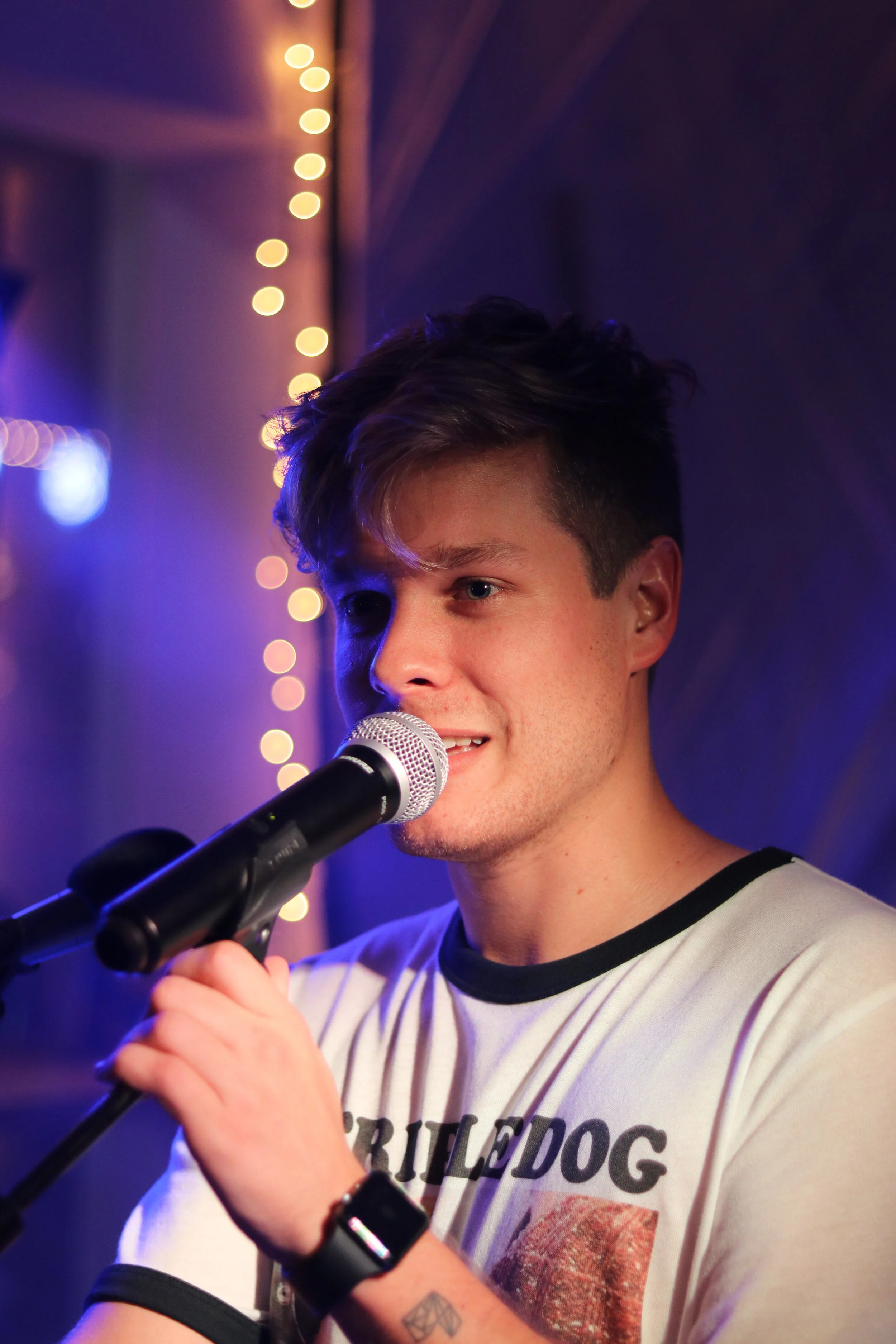A young man with styled hair singing into a microphone on stage, with purple and yellow bokeh lights in the background.