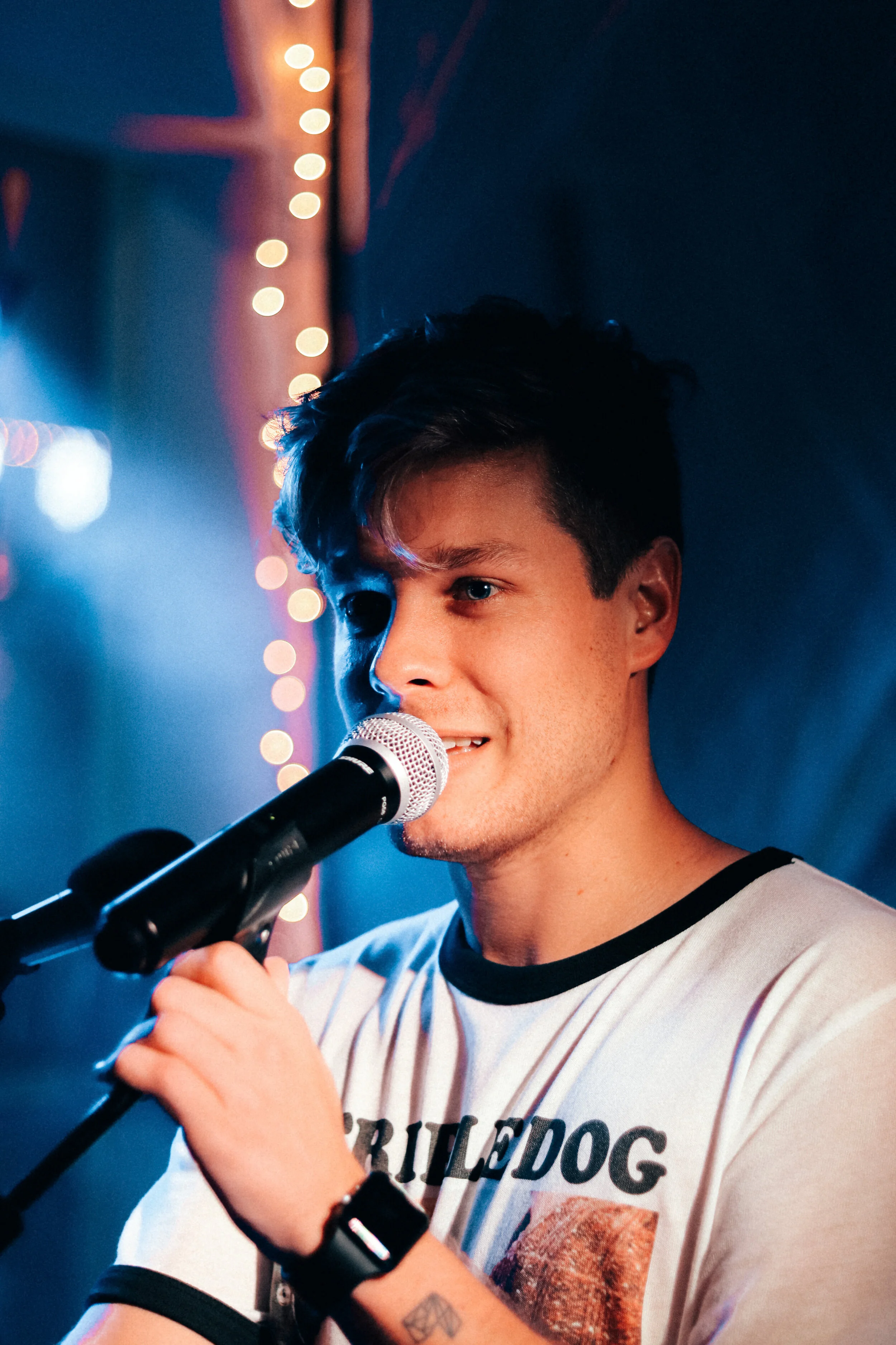 Young man with short dark hair singing into a microphone on stage, with blurred string lights in the background.