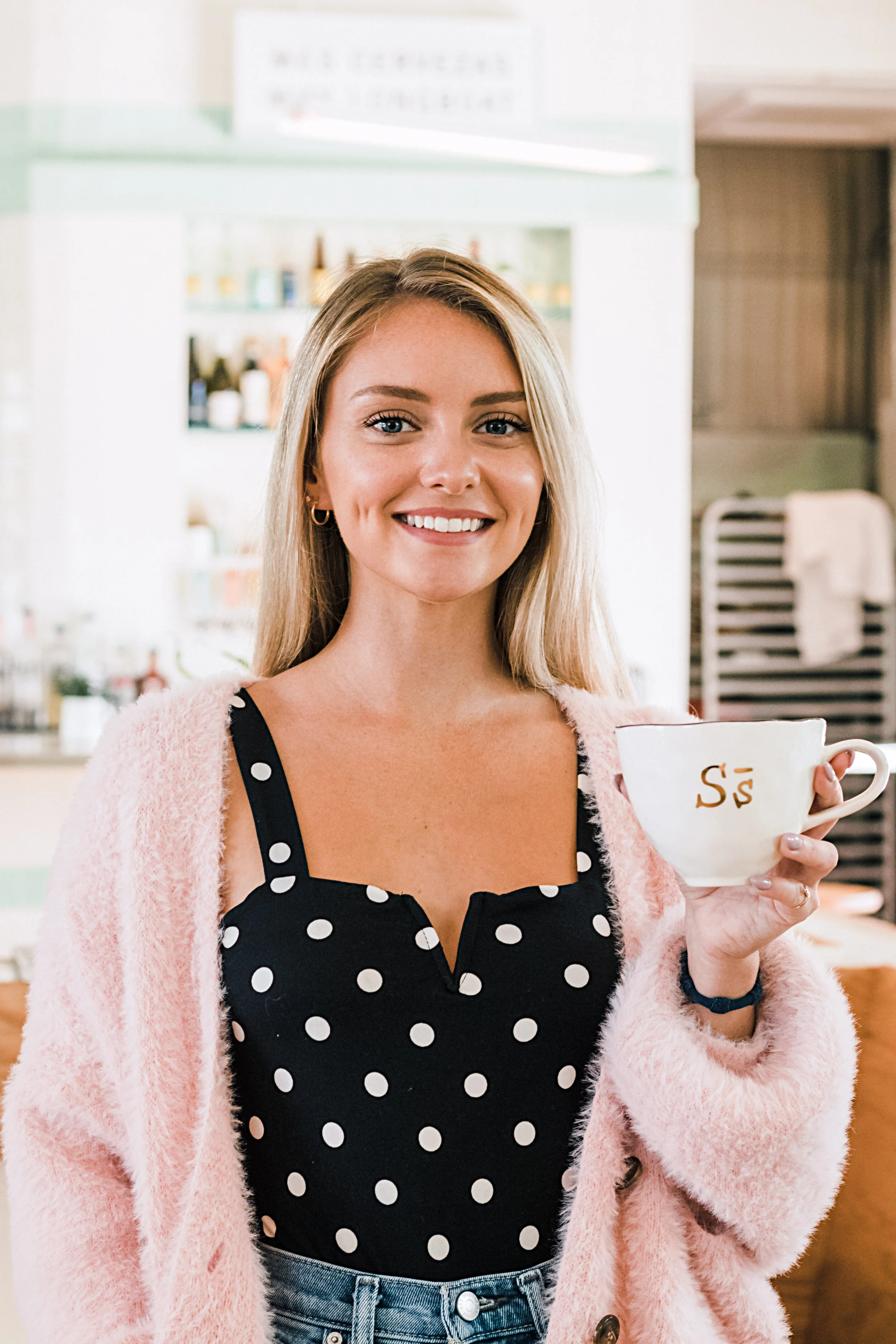 A smiling woman with blonde hair, wearing a black polka dot top and pink fuzzy cardigan, holding a white mug with gold 'S' in a cozy cafe.