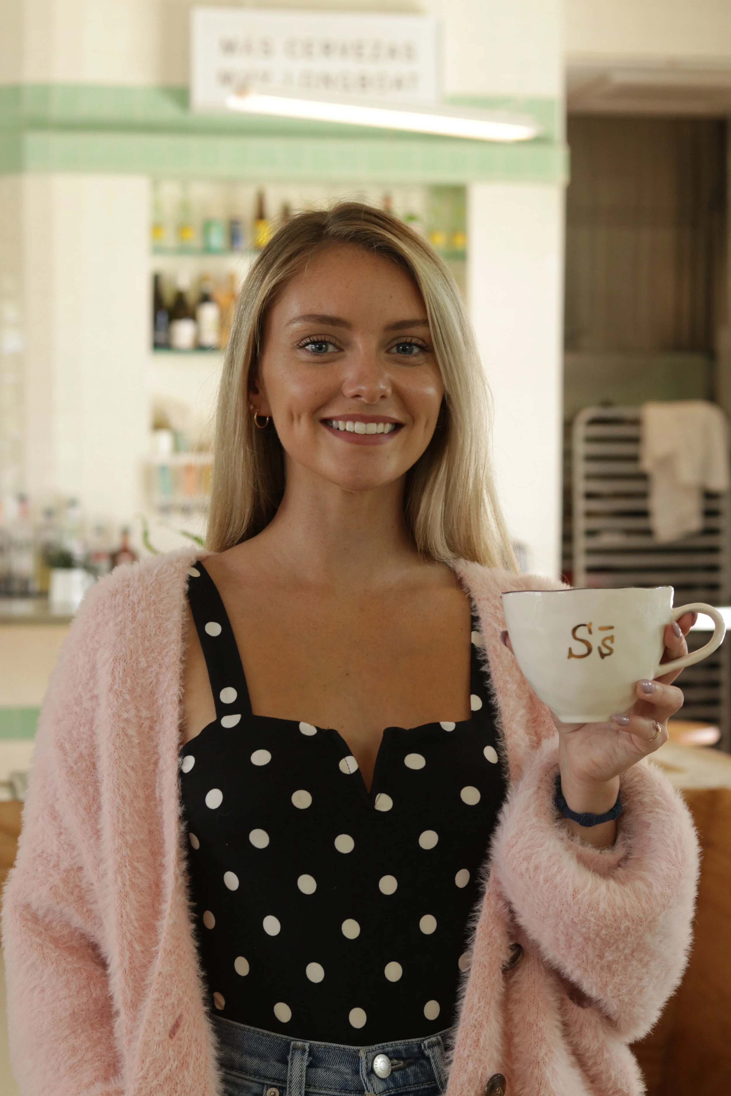 A smiling woman holding a coffee mug with the letter 'S' on it in a cafe setting, wearing a black polka dot top and pink cardigan.