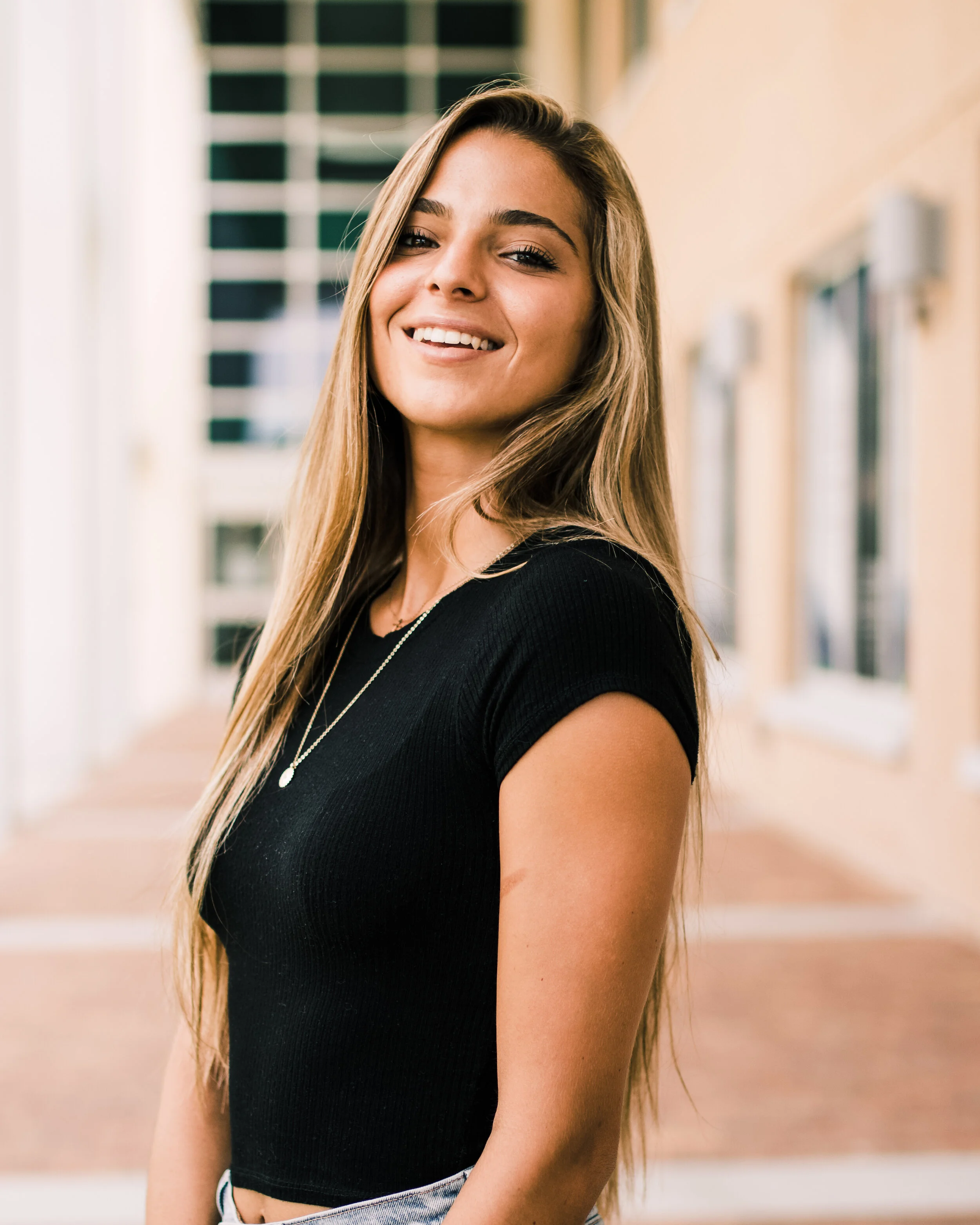 A young woman with long blonde hair, wearing a black short-sleeve top and a necklace, standing outdoors in front of a building with windows and smiling at the camera.