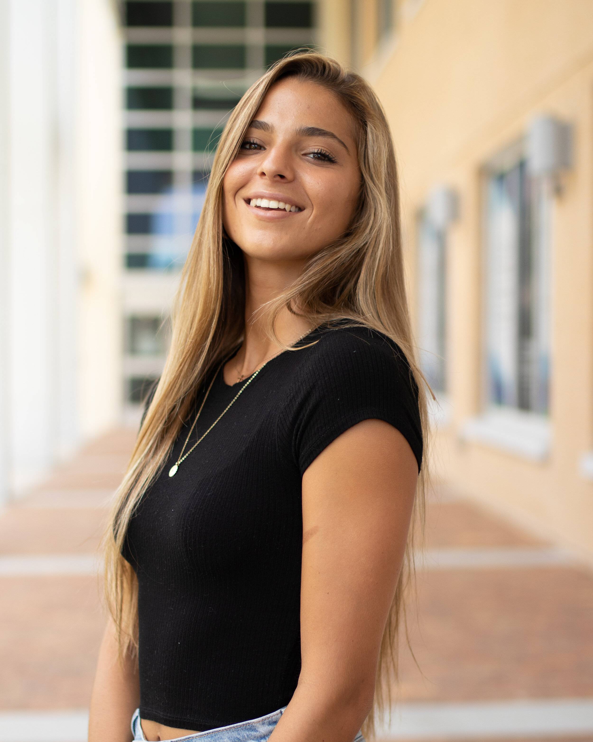 A young woman with long blonde hair smiling, wearing a black top and a necklace, standing outdoors with a background of a building with windows and a walkway.
