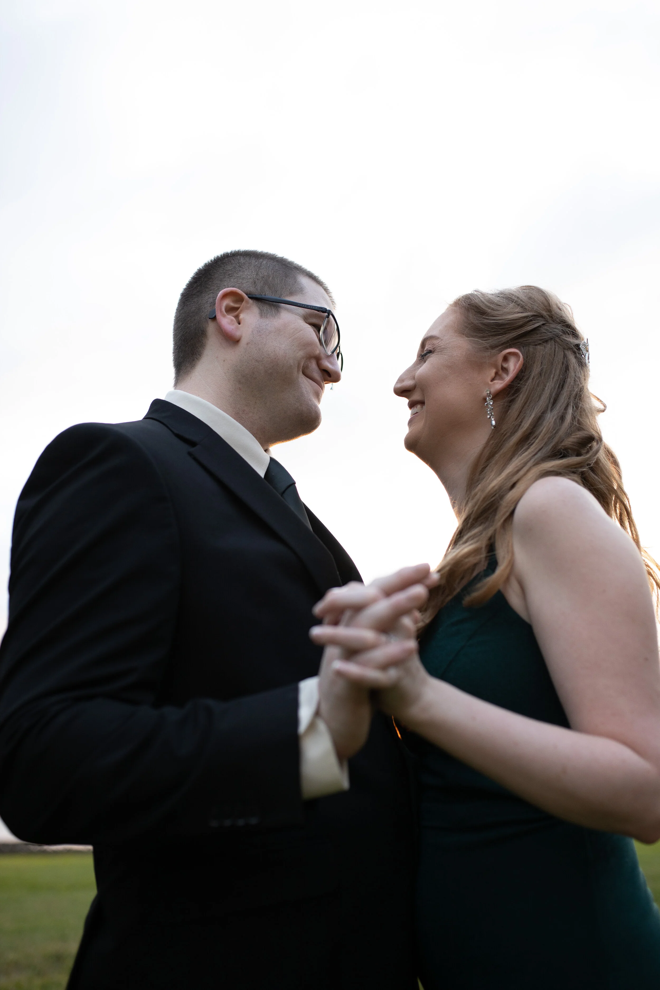 A couple in wedding attire dancing outdoors with bright sky in the background, smiling at each other.