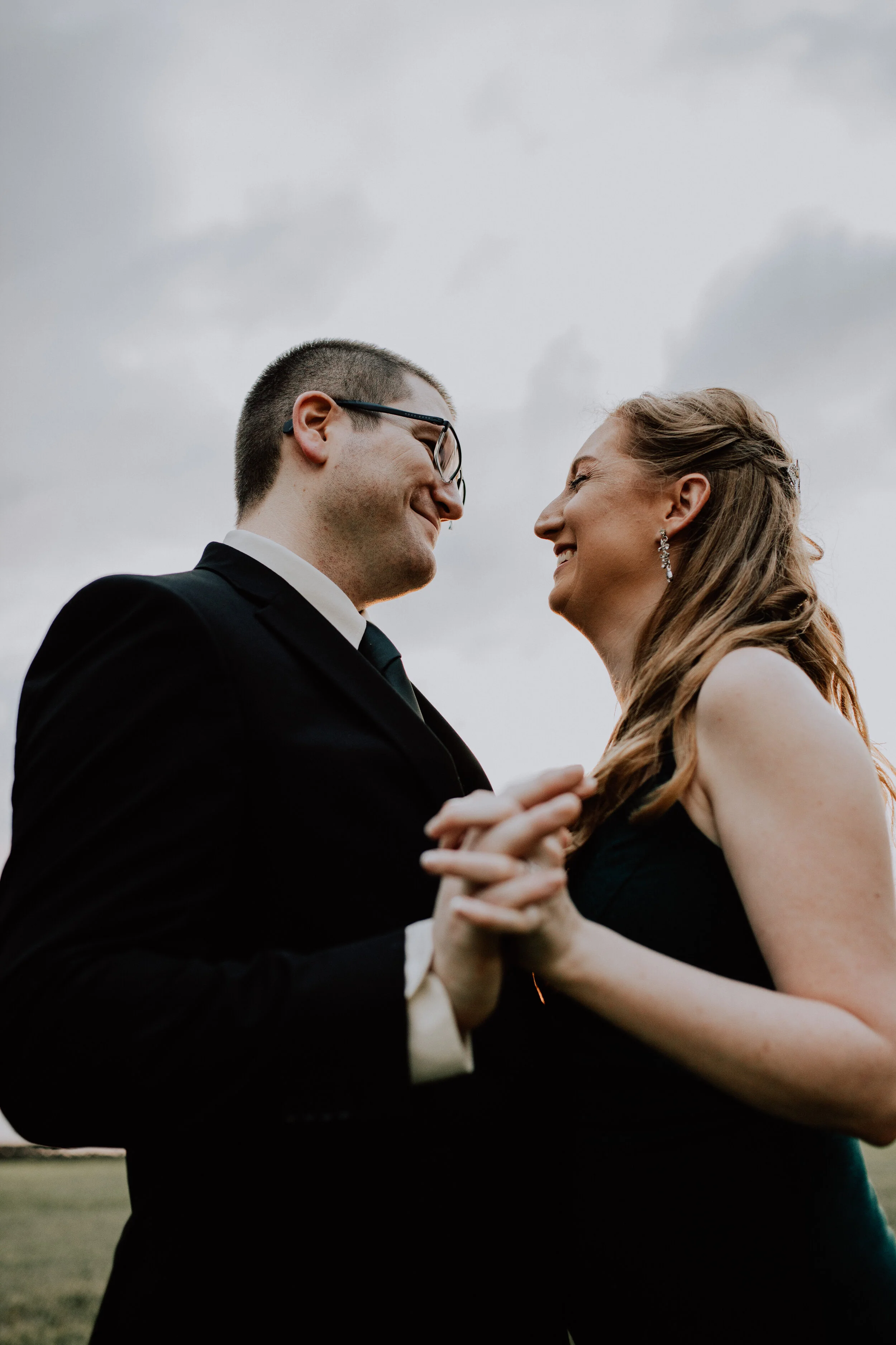 A couple dressed in formal attire, smiling and holding hands outdoors on a cloudy day.