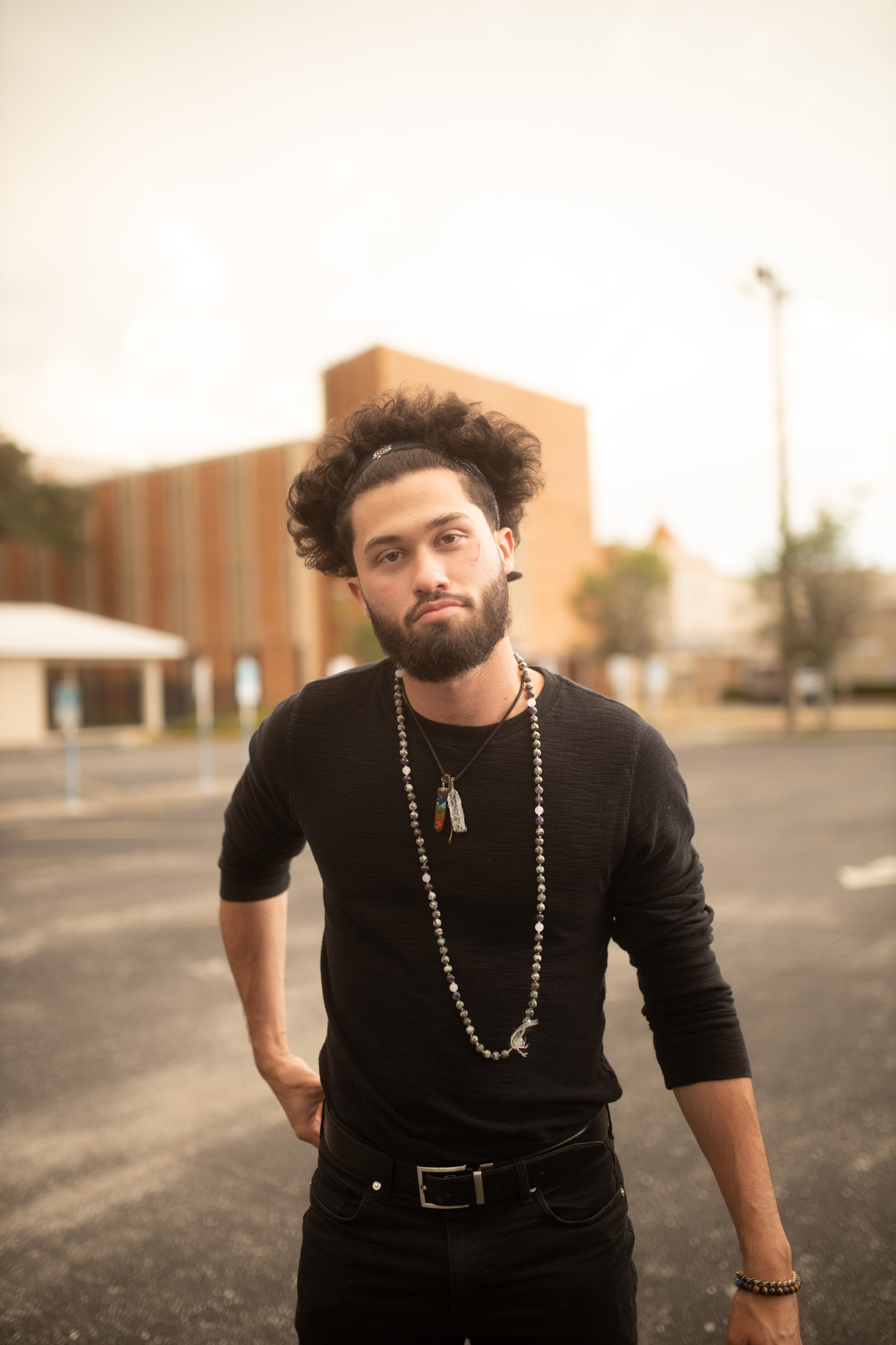 A young man with curly hair and a beard stands in an outdoor parking lot, wearing a black long-sleeved shirt, bracelets, and necklaces, with a slightly serious expression.