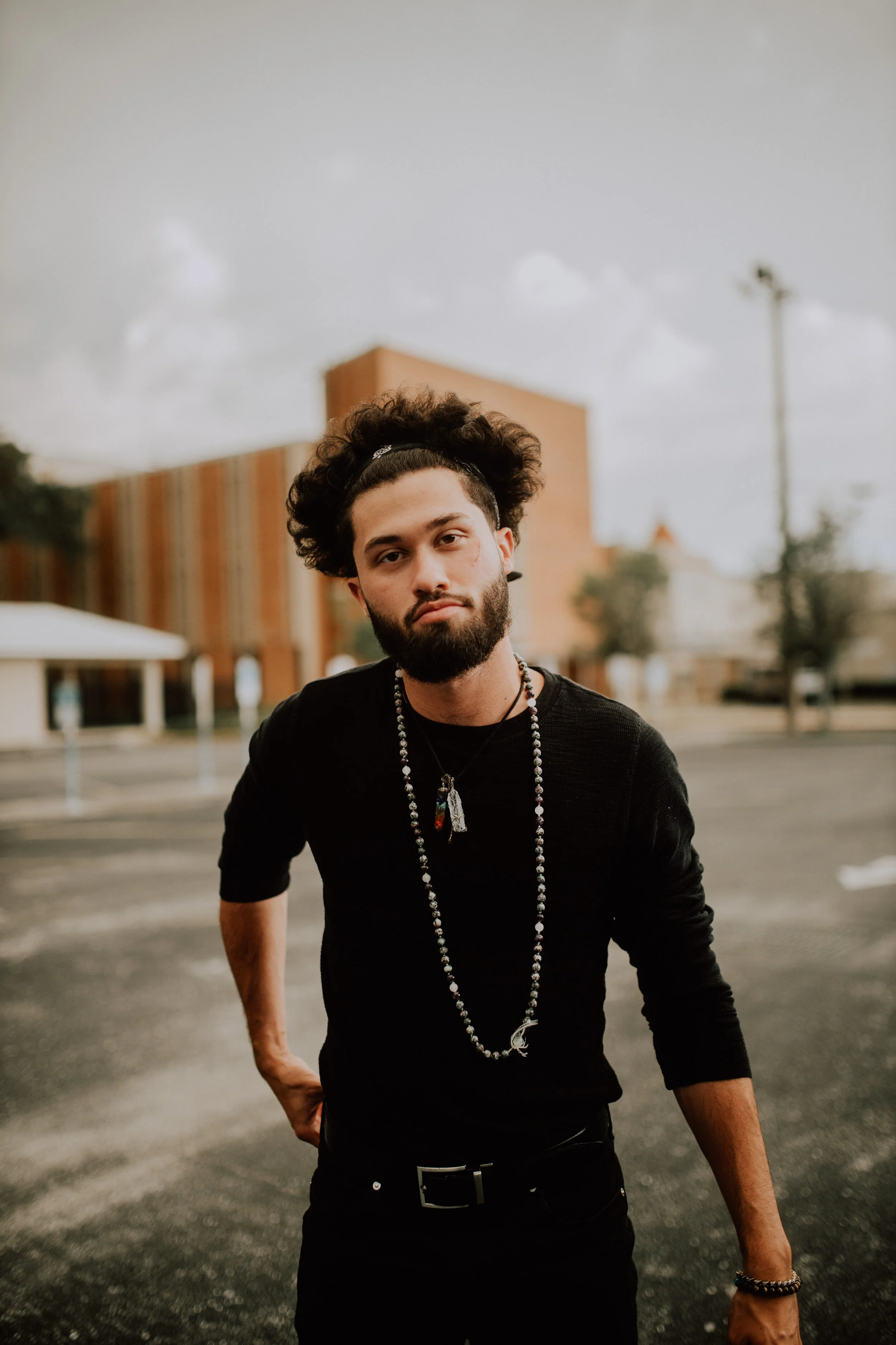 A young man with curly hair and a beard wearing a black shirt with jewelry poses give a serious expression outdoors.