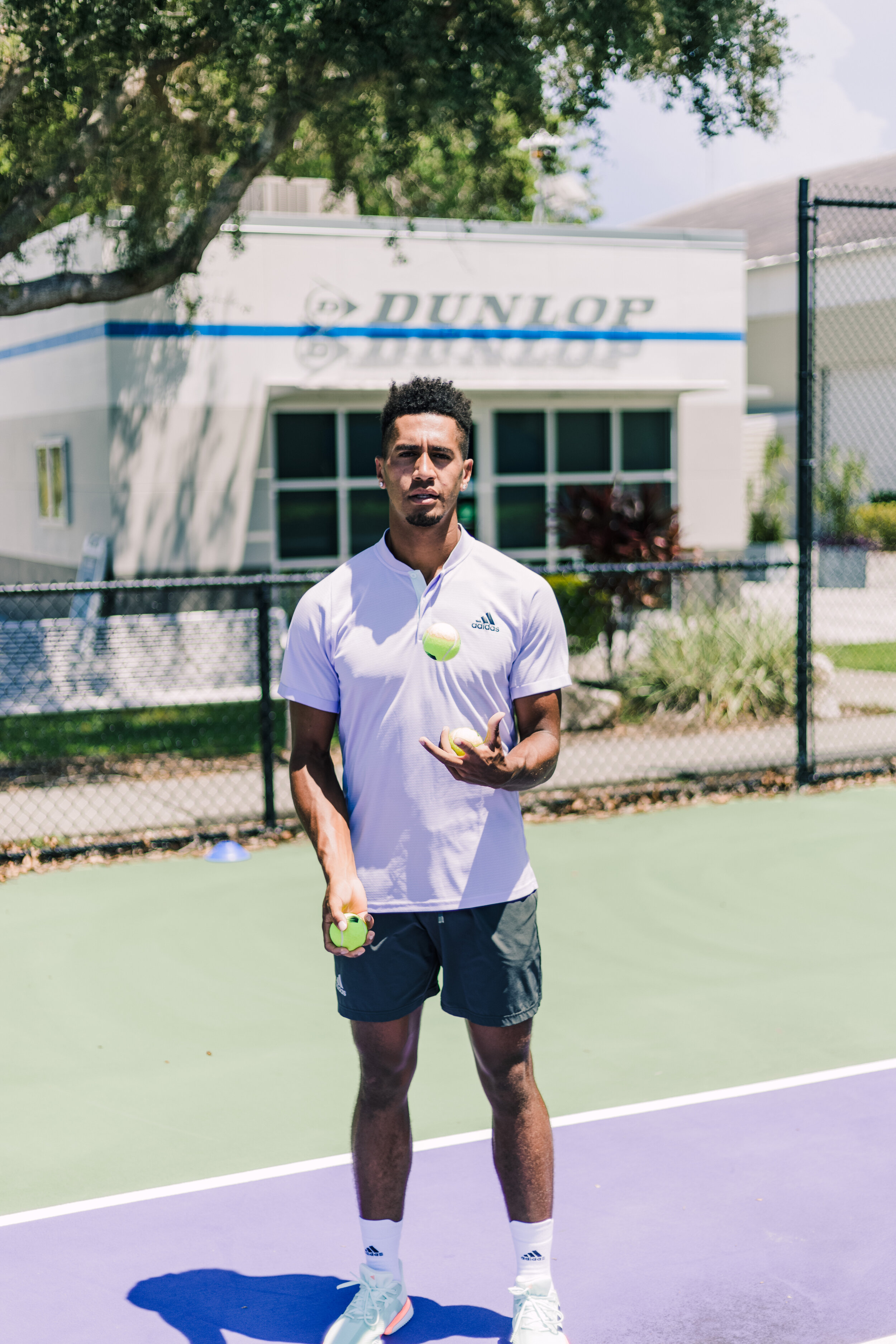 Man standing on tennis court holding tennis balls, wearing white athletic shirt and black shorts, with a fence and building labeled 'Dunlop' in the background.