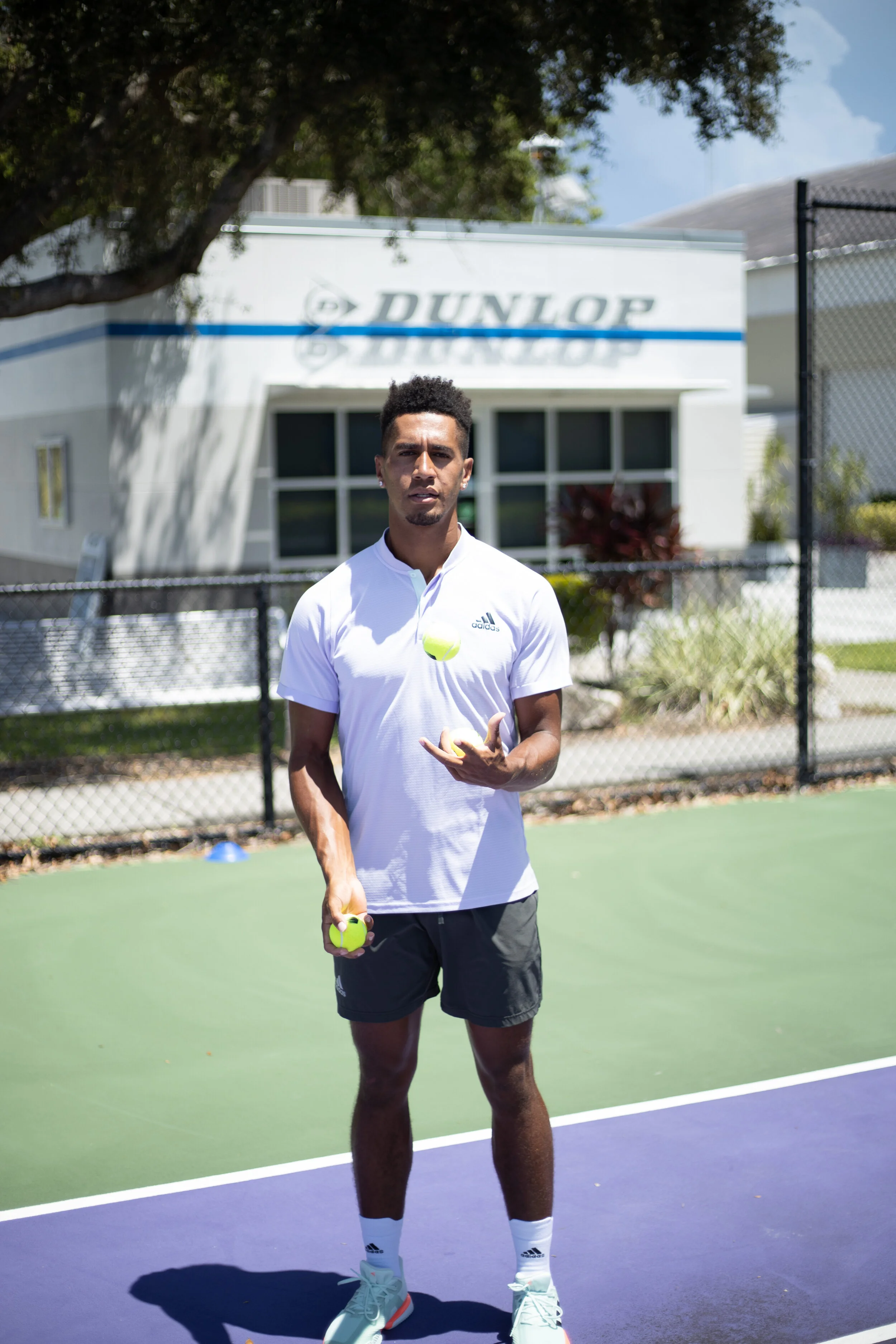 A young man standing on a tennis court, holding tennis balls, with a Dunlop sign and a building in the background.