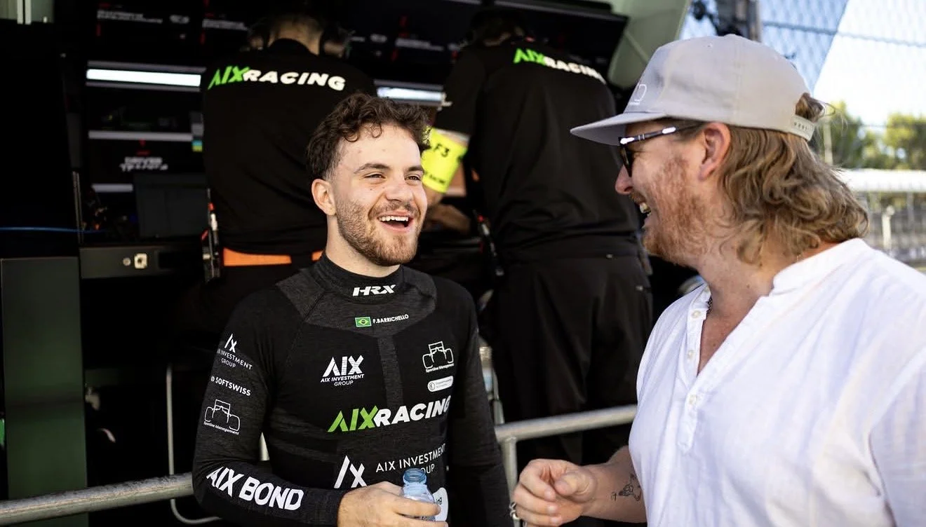 A young race car driver with a beard and wearing a black racing suit with sponsor logos, smiling and holding a water bottle, converses with an older man wearing a white shirt, sunglasses, and a light gray cap, at a racing event, with team members working in the background.