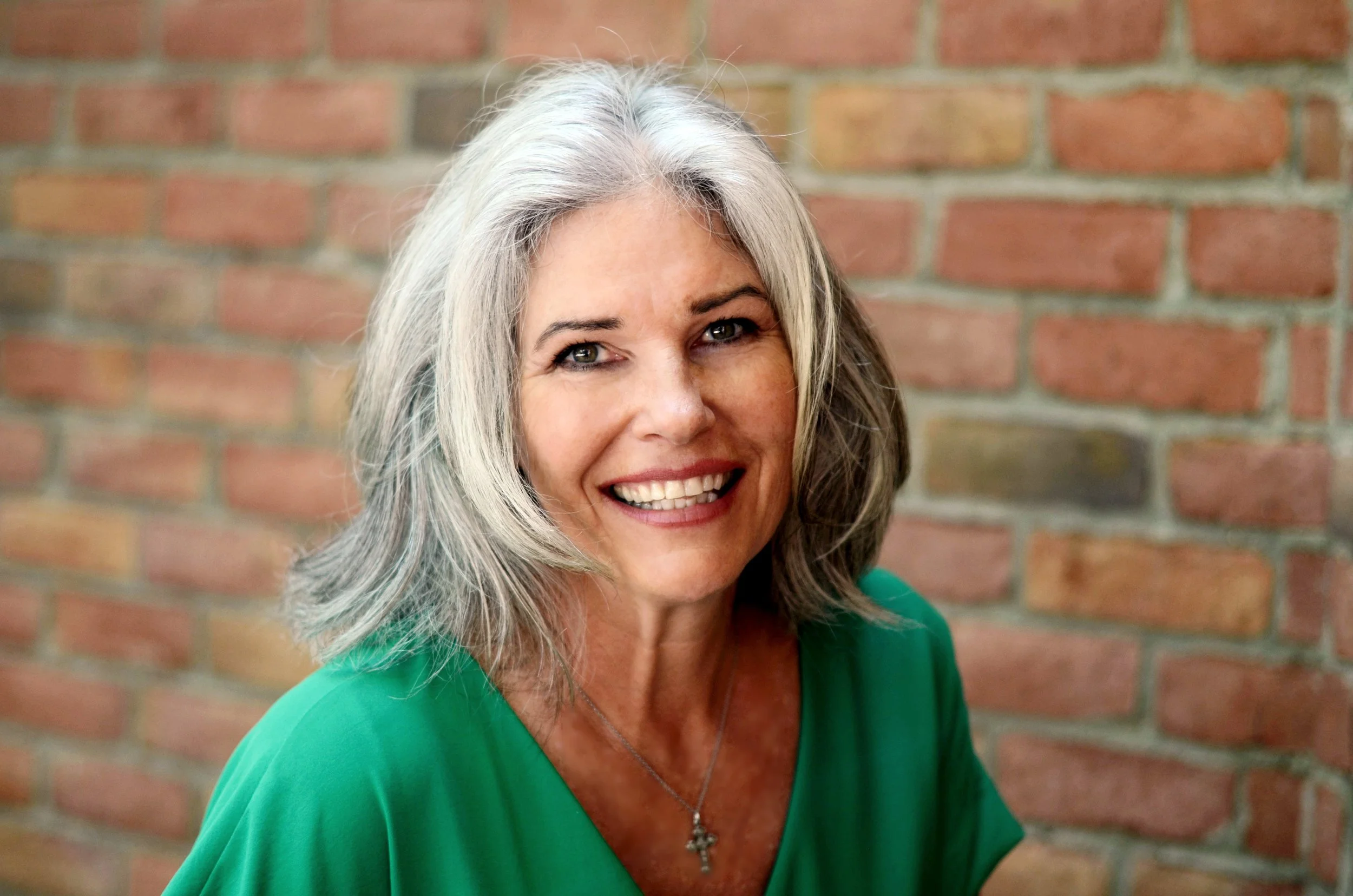 A smiling middle-aged woman with shoulder-length gray hair wearing a green top and a silver cross necklace, standing in front of a brick wall.