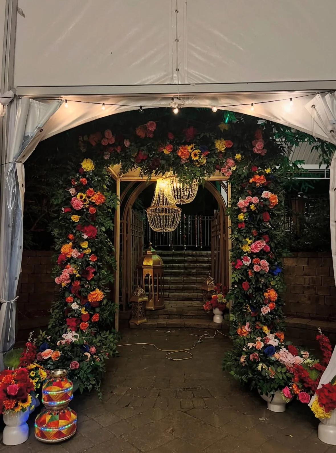 A decorated entrance with an archway covered in multicolored flowers, string lights, and hanging chandeliers. Lanterns and flower arrangements are placed along the sides of the path leading to steps.