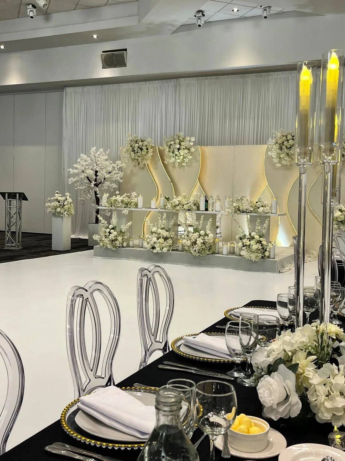 Elegant wedding reception setup with a white and black color scheme, floral arrangements, table settings with white napkins, glassware, and decorative candles, and a stage decorated with flowers and ambient lighting.