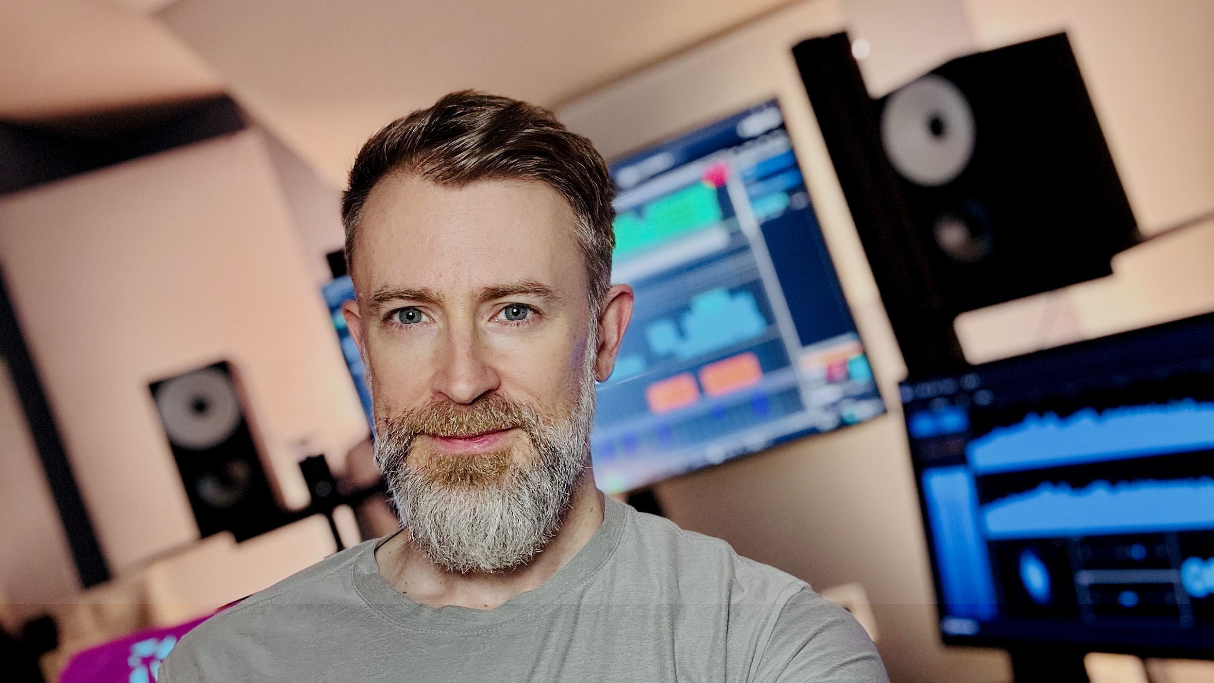 A man with a beard and short dark hair taking a selfie in a music or recording studio with multiple audio monitors and computer screens displaying mixing software