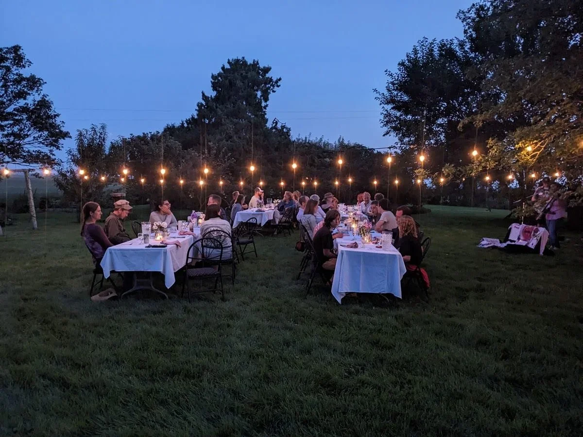 Outdoor dinner party at dusk with long tables covered in white tablecloths, string lights hanging overhead, and guests seated eating and talking in a grassy area surrounded by trees.