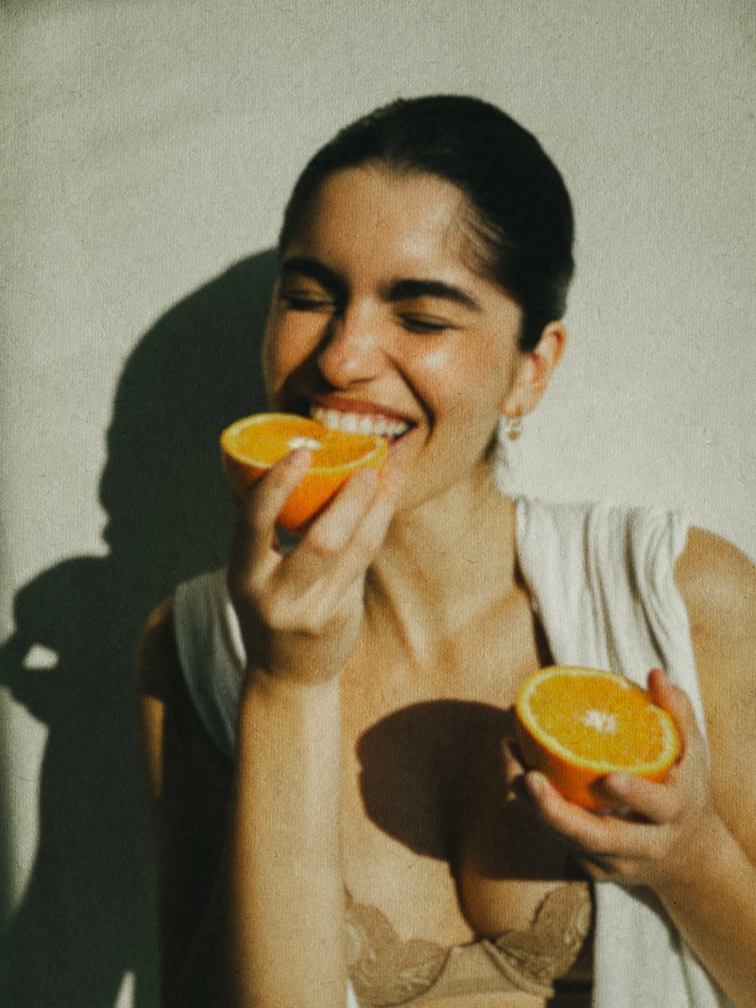 A woman with dark hair, smiling with eyes closed, holding half an orange close to her mouth and another half in her other hand.