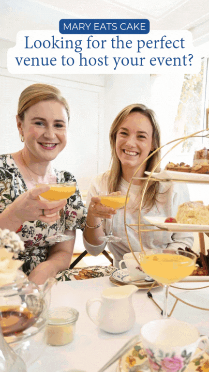 Two women enjoying a tea party with desserts, holding glasses of orange juice, seated at a table with various cakes and pastries, during a daytime event.