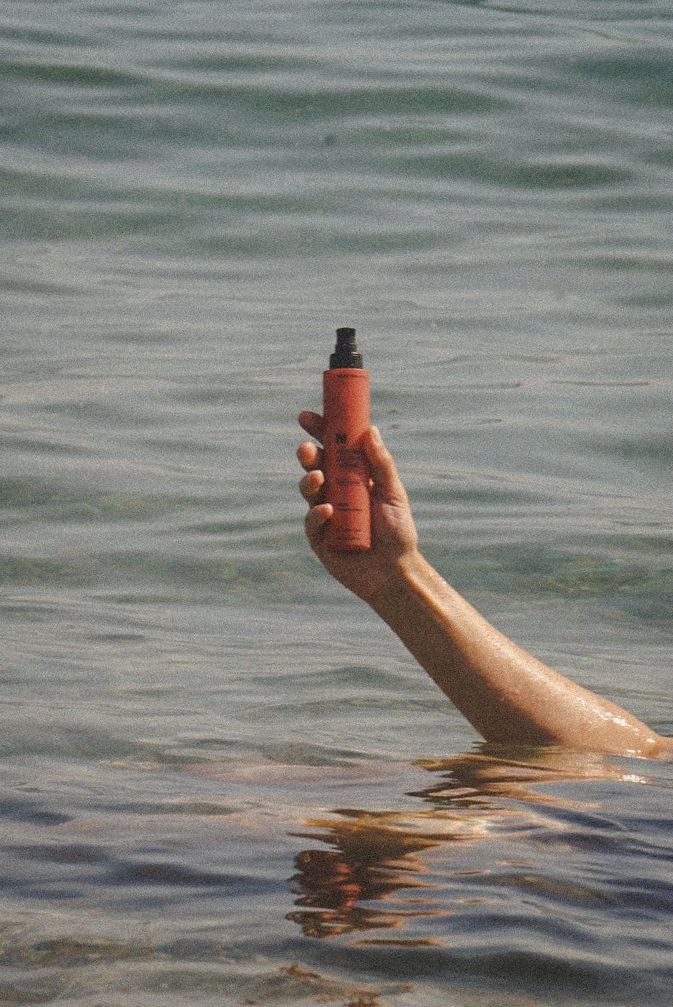 A person's arm extending out of the water, holding a red spray bottle with a black nozzle, in a body of water with gentle ripples.