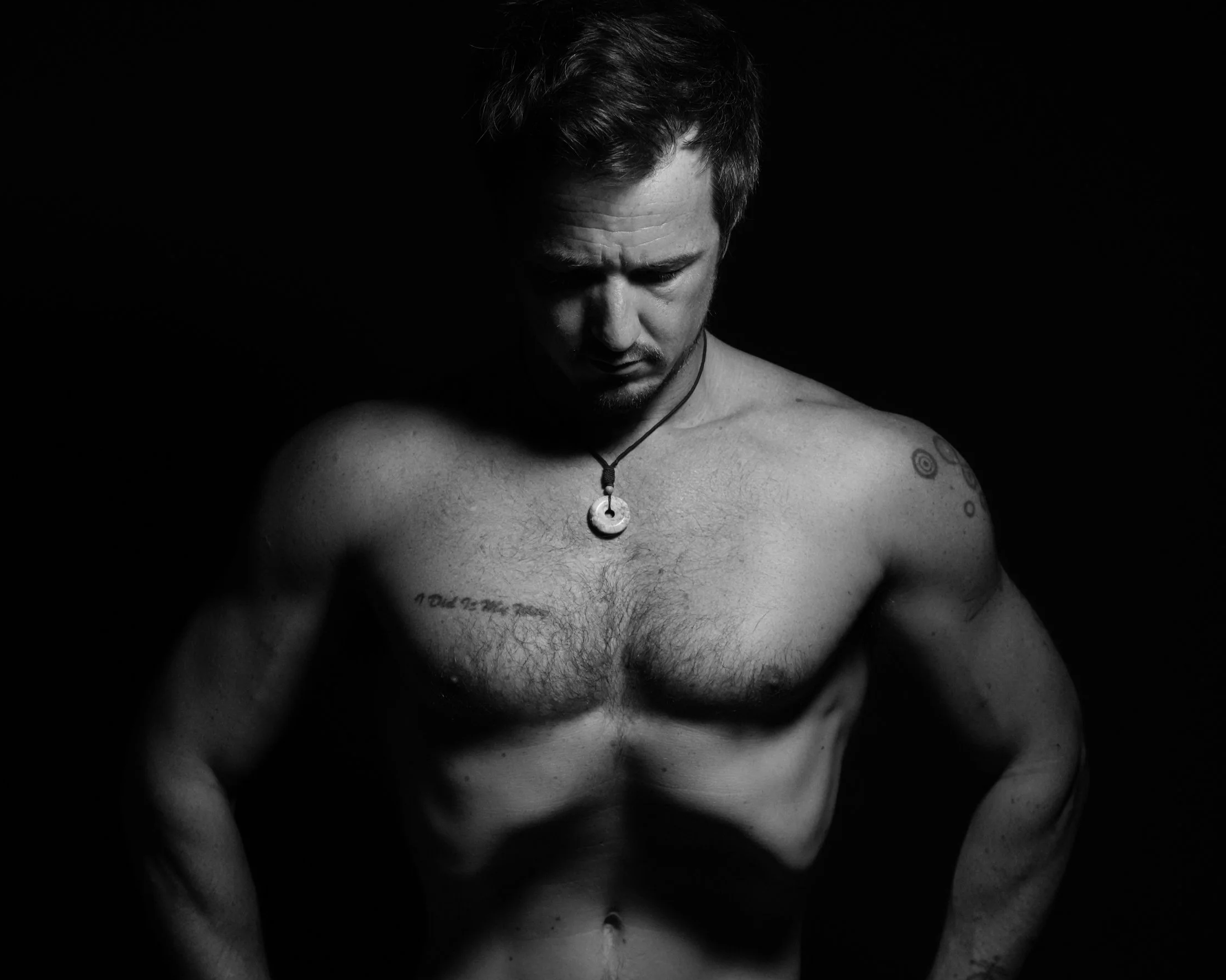 Black and white photograph of a shirtless man looking down with a contemplative expression, wearing a necklace with a circular pendant, with tattoos on his chest and arms, against a dark background.