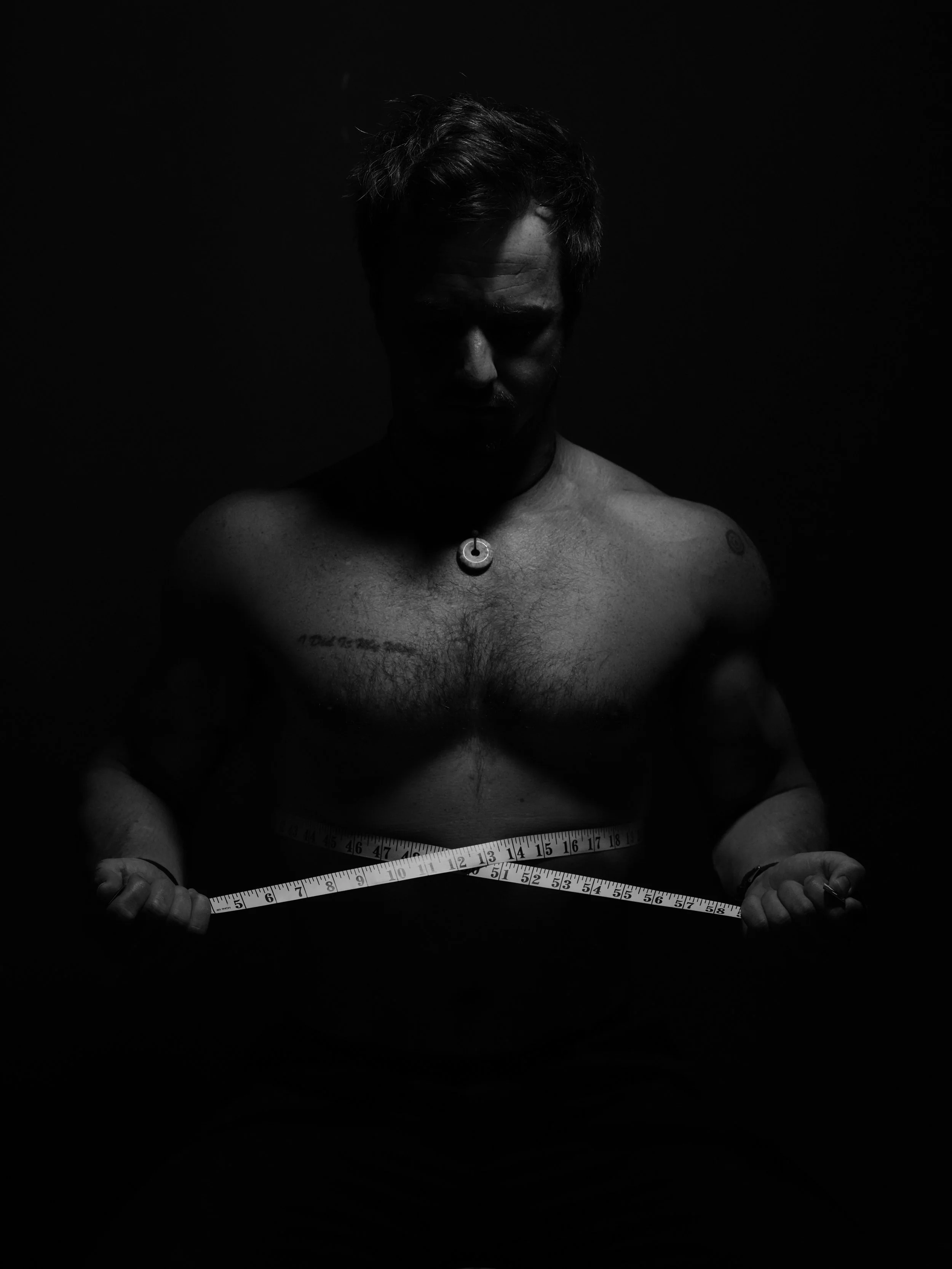A black and white photo of a shirtless man with tattoos, holding a measuring tape around his waist in a dark setting with shadowed lighting.