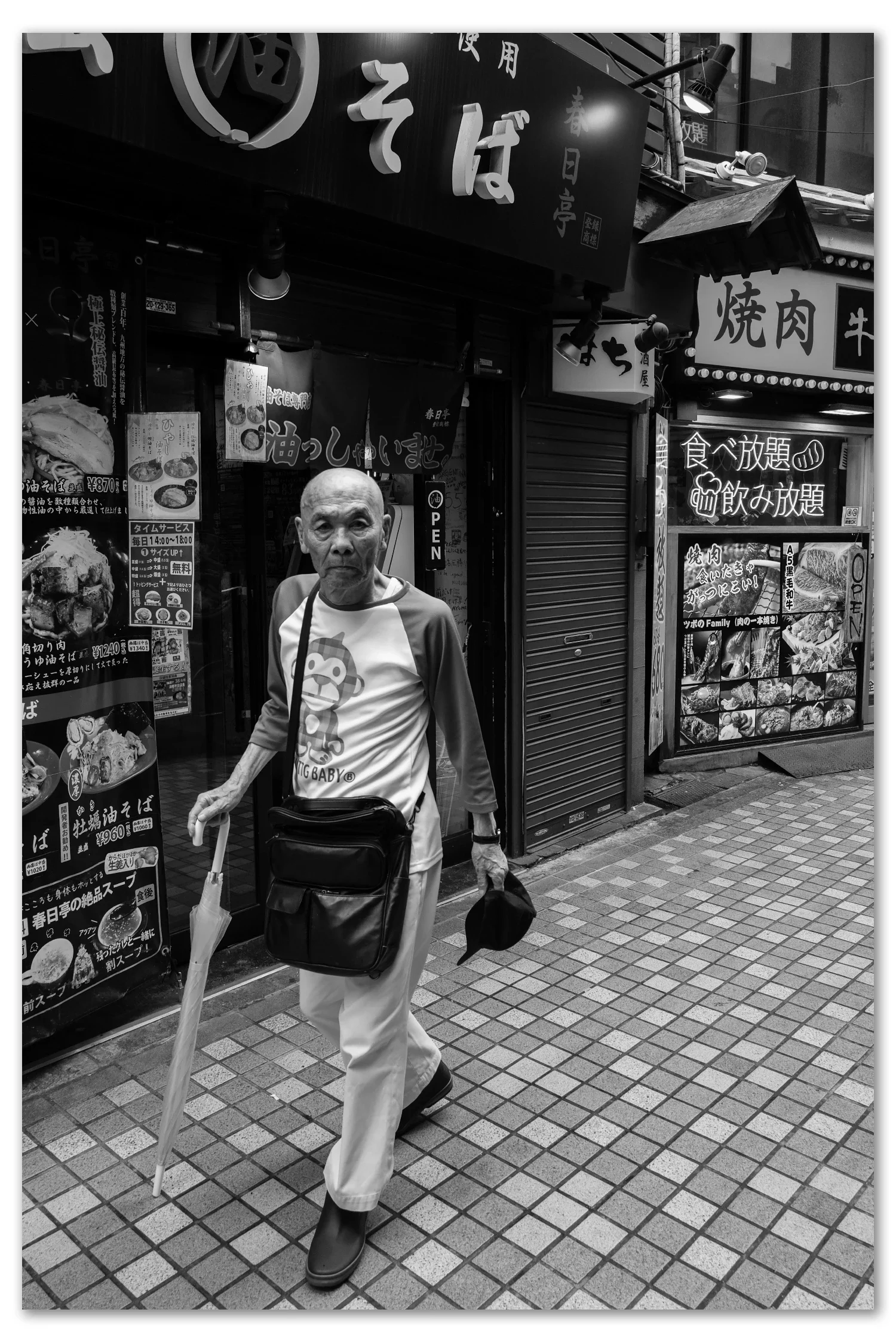 Black and white street photograph, Nakano, Tokyo — an older man walks down a soba alley using his umbrella as a cane, Leica Q2 Monochrom 28mm