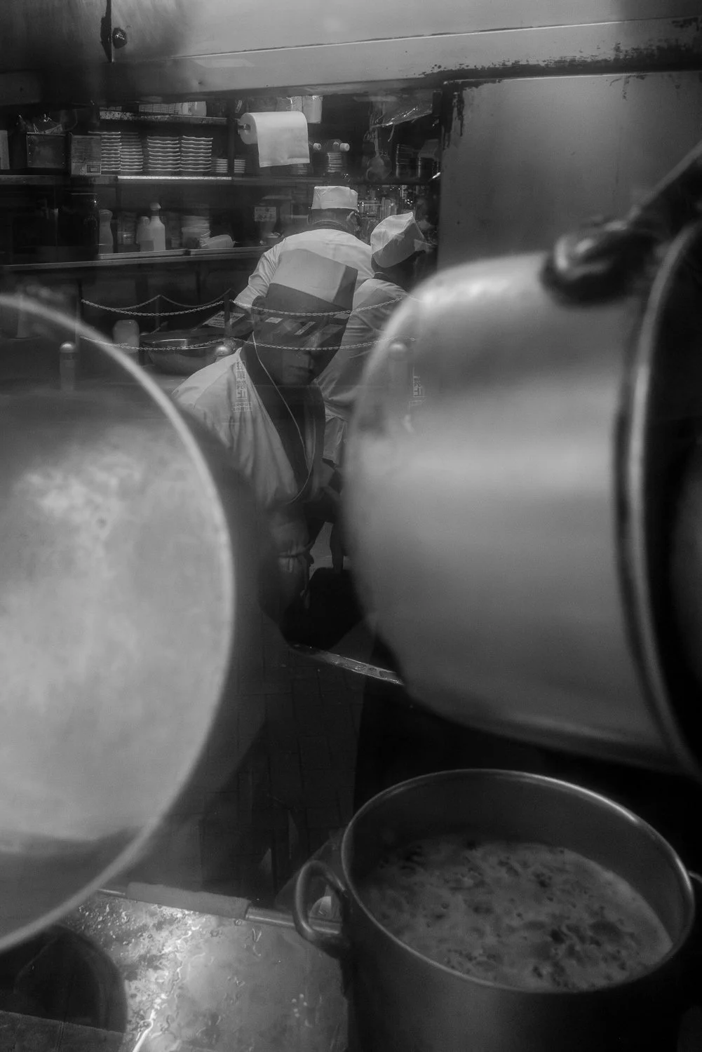 Cooks in white caps work behind stacked stockpots, seen through a smeared shop window, Shibuya