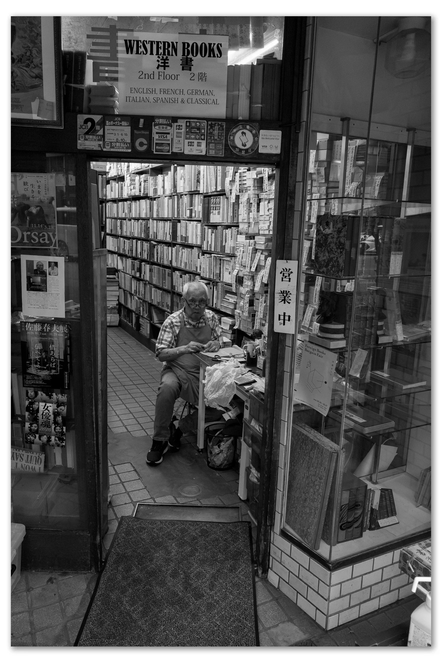 A man seated deep inside a narrow Western Books shop stacked floor to ceiling, framed by the doorway, Jimbocho