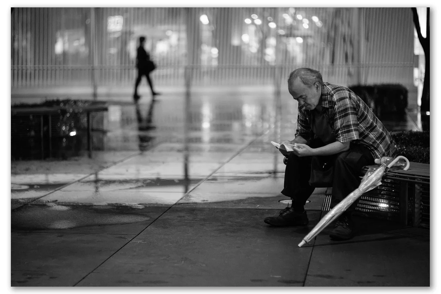 A man in a checked shirt reads on a bench in the rain-soaked plaza of the Tokyo International Forum, a closed umbrella at his feet]
