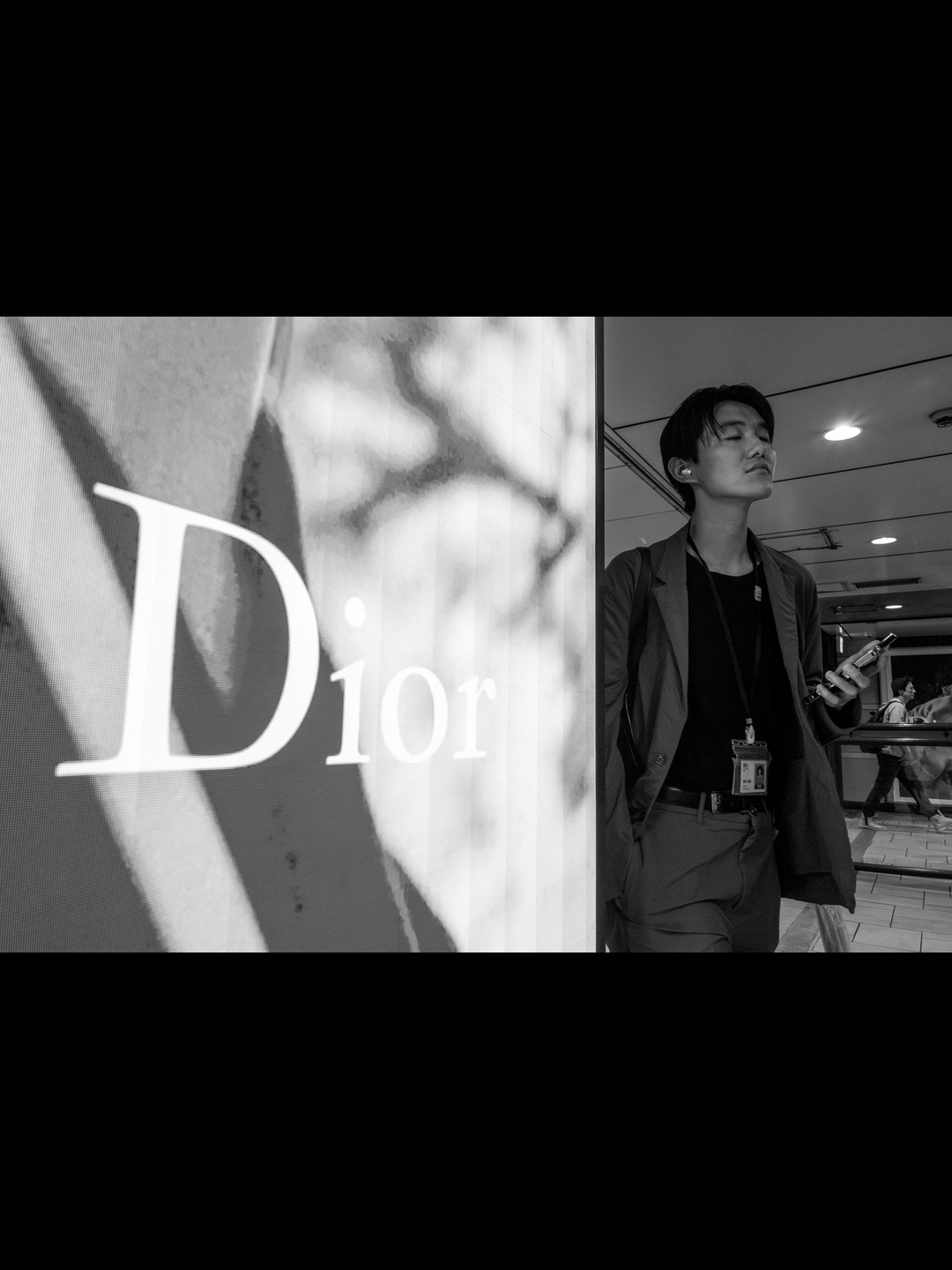 A man in a suit walks past a Dior light box in an underground concourse in Shibuya, eyes closed, earbud in, forged at 28mm on a Leica Q2 Monochrom