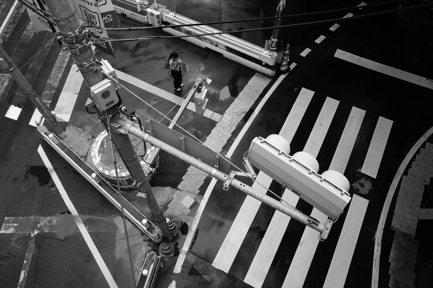 Looking straight down from a pedestrian bridge at a traffic signal, a zebra crossing and one small figure waiting on the wet corner below, Shibuya