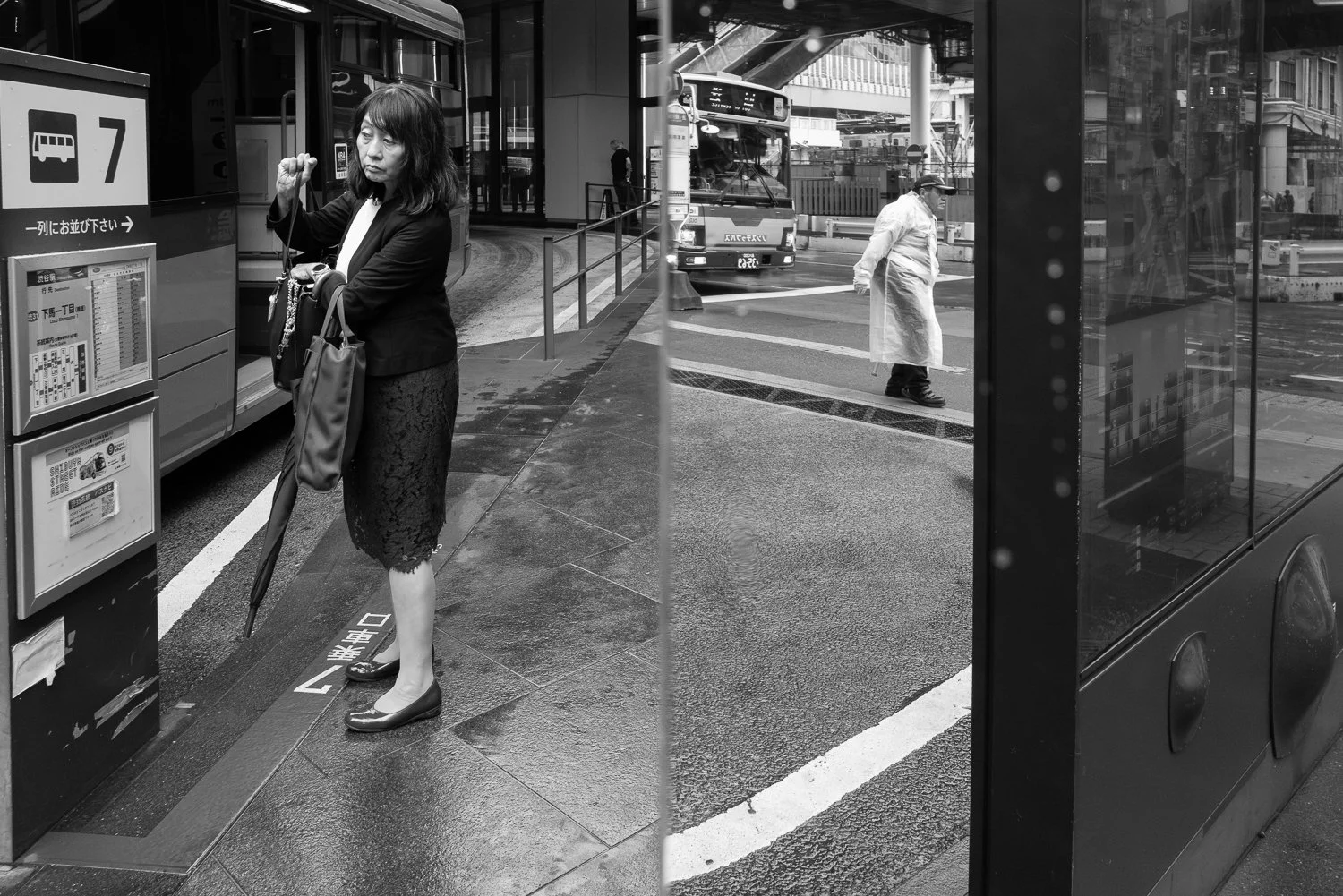 A woman at bus stop number 7 fastens her closed umbrella while a traffic guard in a clear raincoat crosses behind her, Shibuya