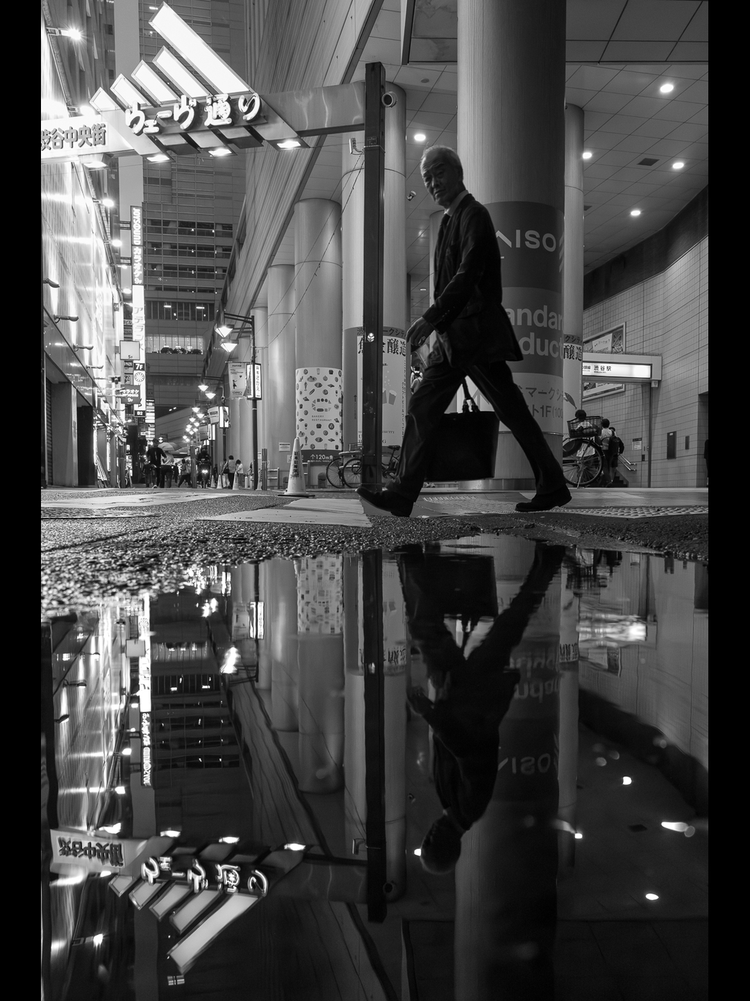 A man in a suit crosses Wave-dōri in Shibuya Chūō-gai at dusk, his reflection upside down in a rain puddle, a Shibuya Mark City West sign on the pillar behind, forged at 28mm