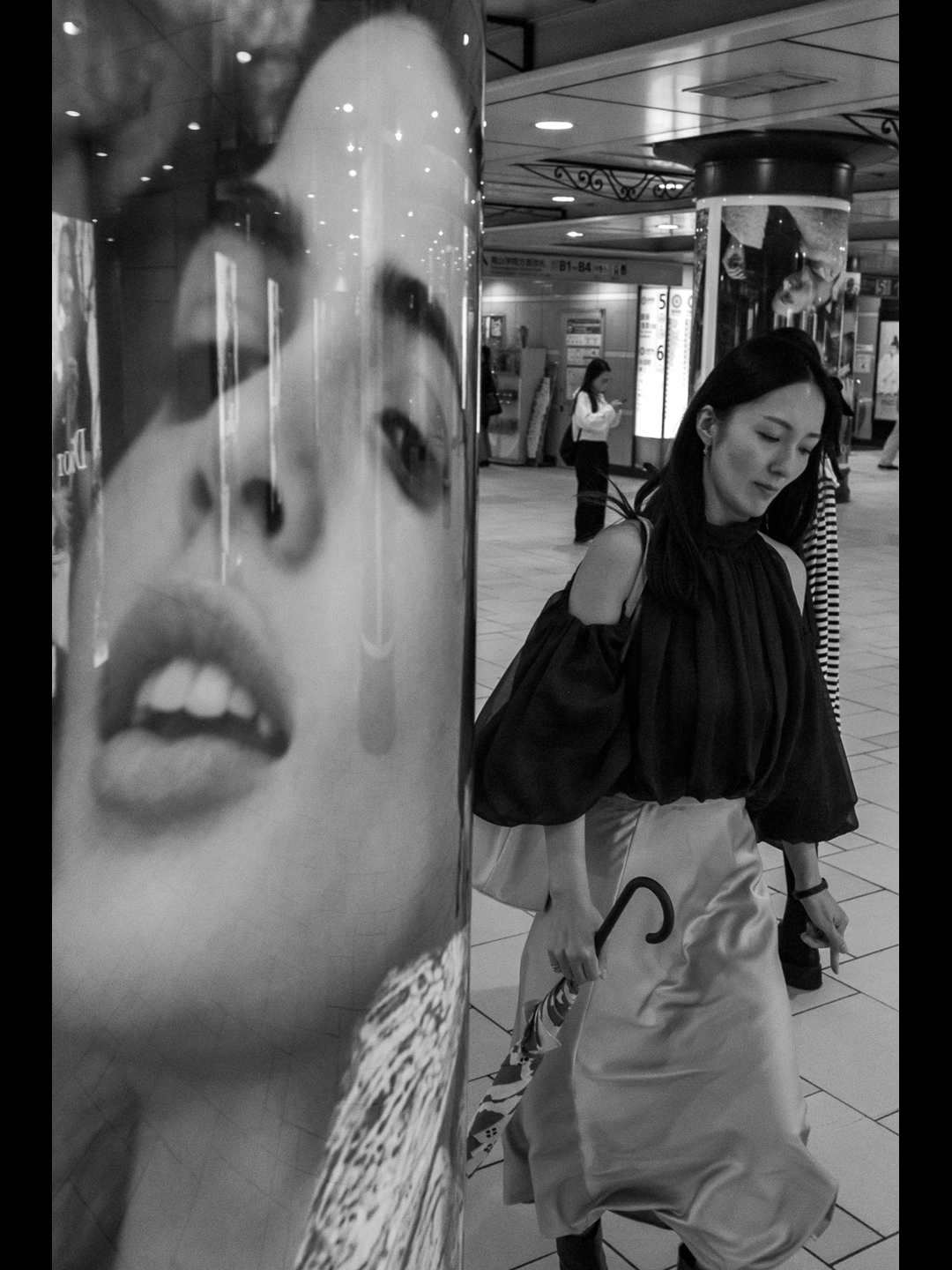 A woman walks past a column wrapped in a giant face in a Shibuya underground concourse, umbrella hooked over her wrist, forged at 28mm on a Leica Q2 Monochrom