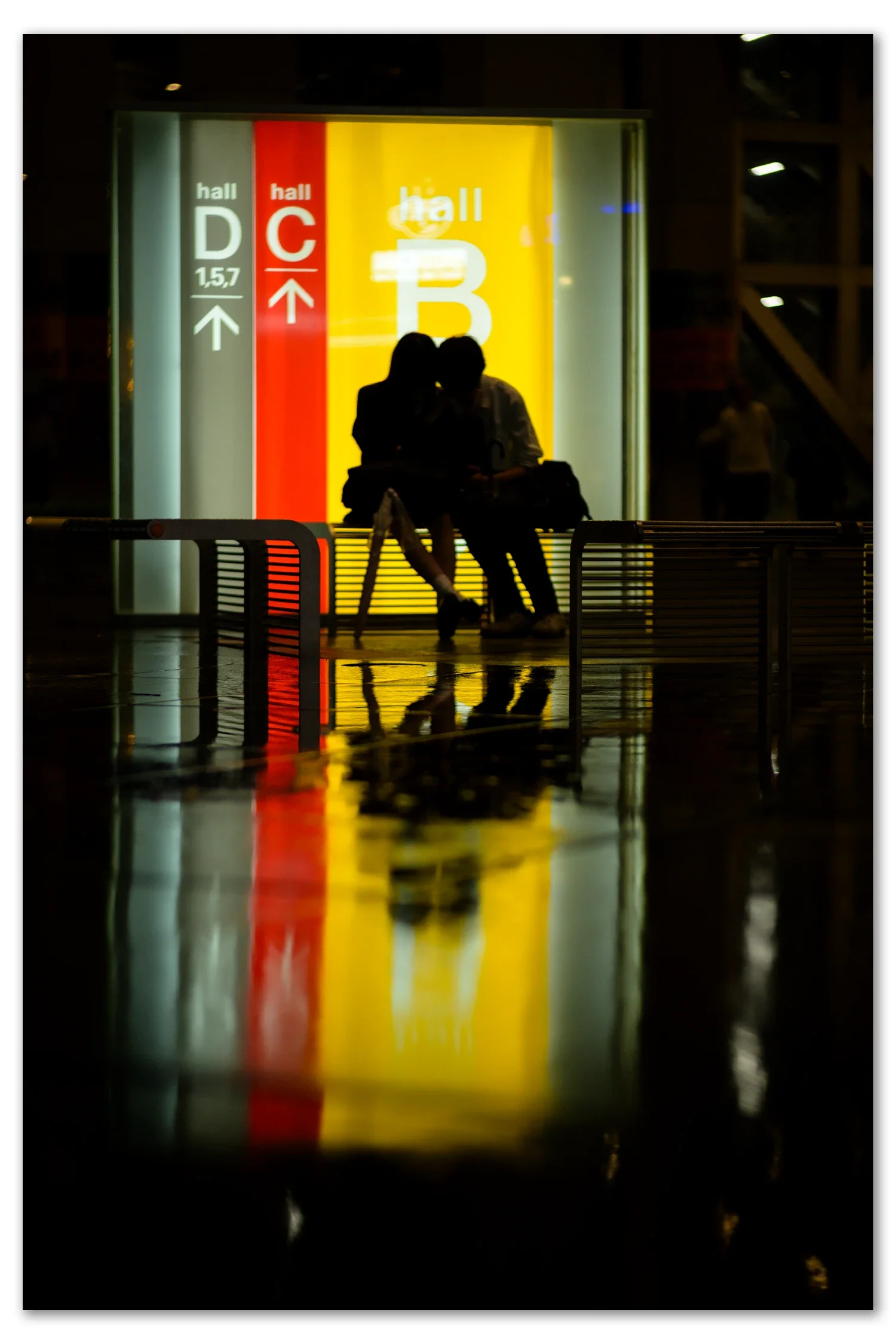 A couple silhouetted against the yellow and red Hall B, C and D signage at the Tokyo International Forum, reflected in the wet floor