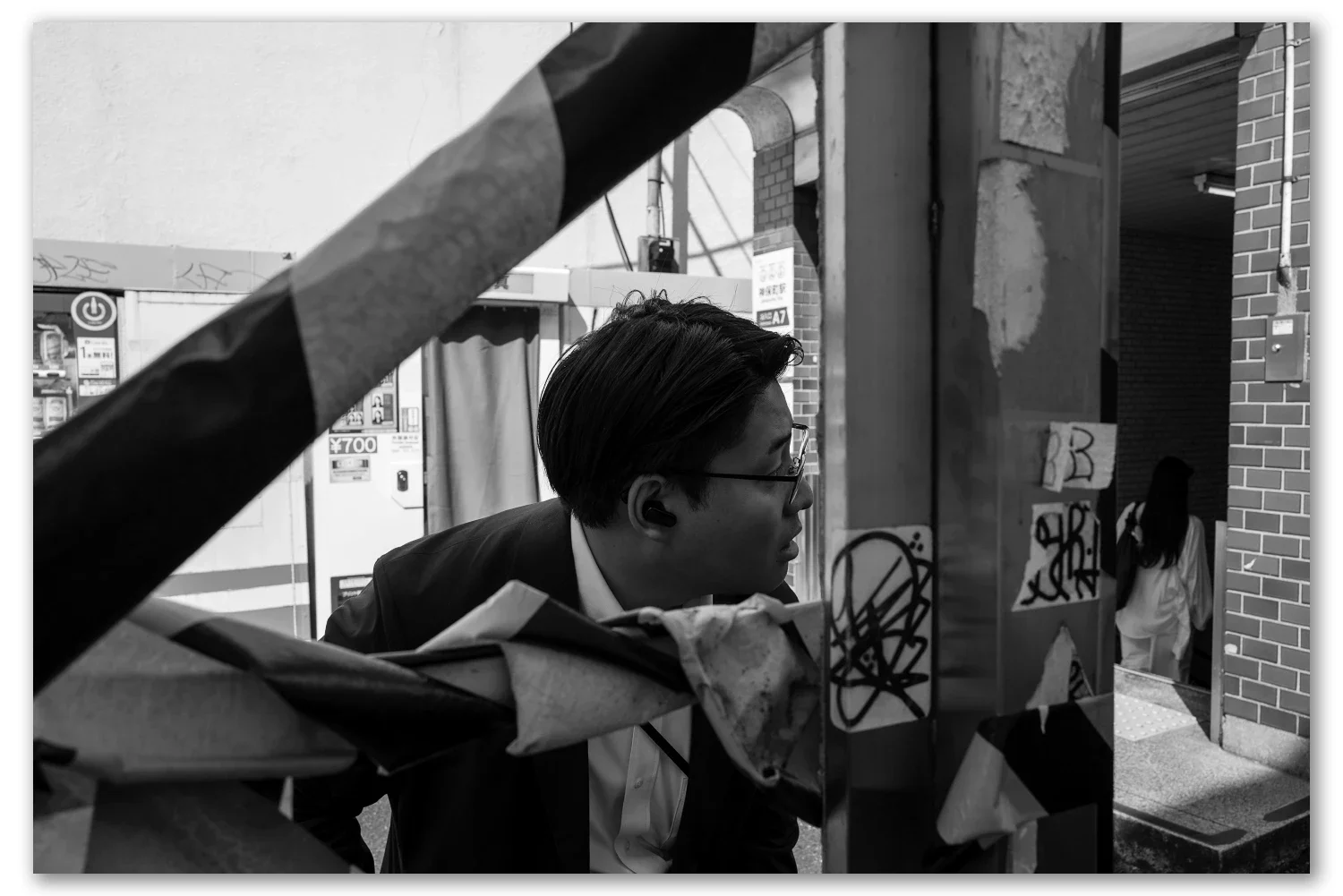A man in a suit glances back through a gap in torn striped barrier tape, vending machines behind him, Jimbocho