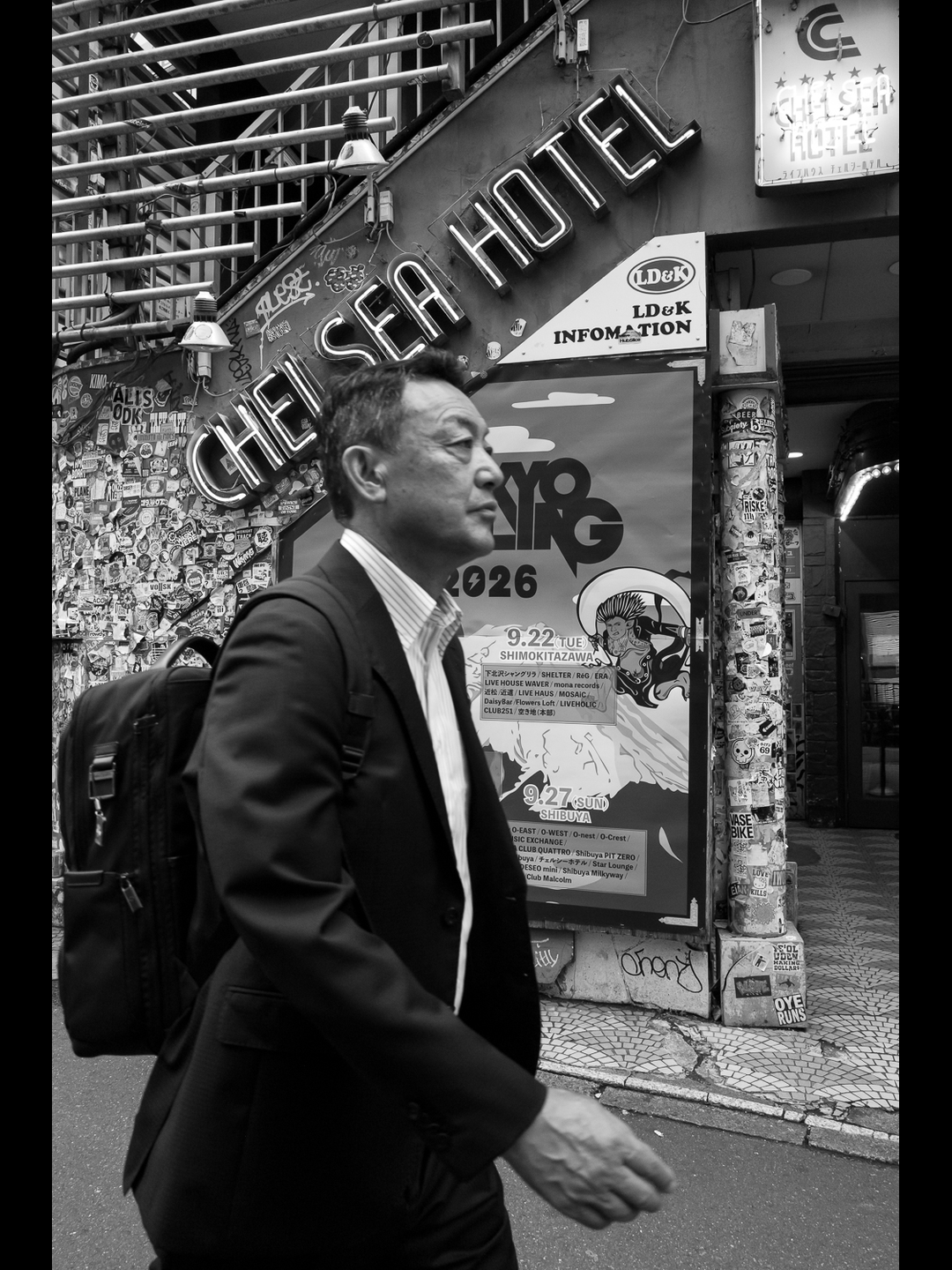 A businessman walks past the neon sign of the Chelsea Hotel live house in Shibuya, a sticker-covered wall and a TOKYO CALLING 2026 poster behind him, forged at 28mm close range