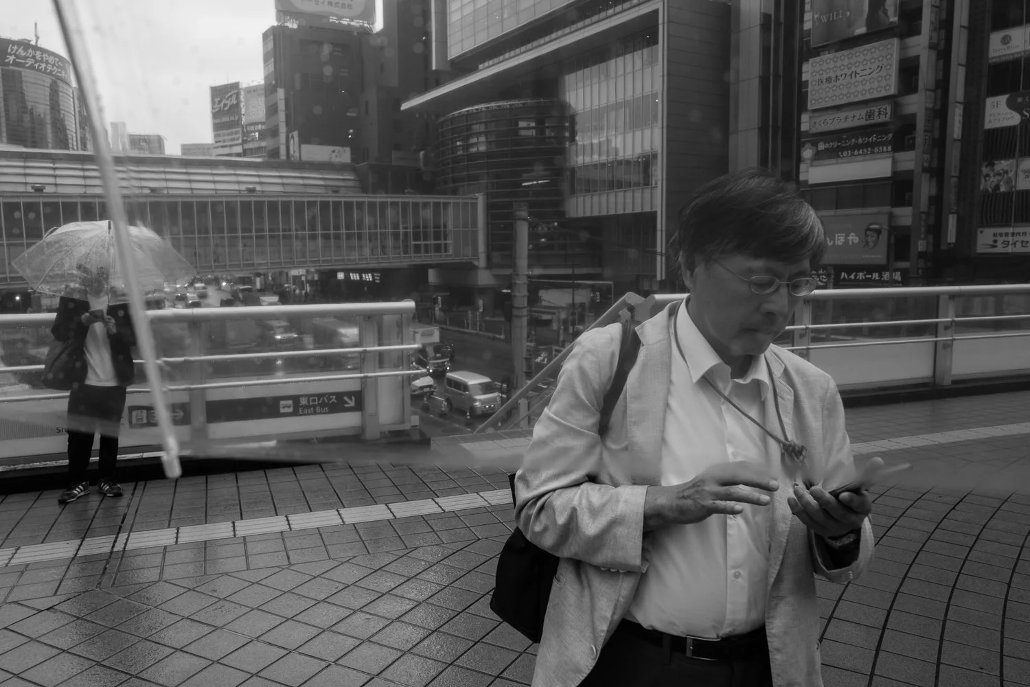 A man in a light jacket reads his phone on a rainy pedestrian deck at Shibuya Station's east exit, the bus terminal and a clear umbrella behind him