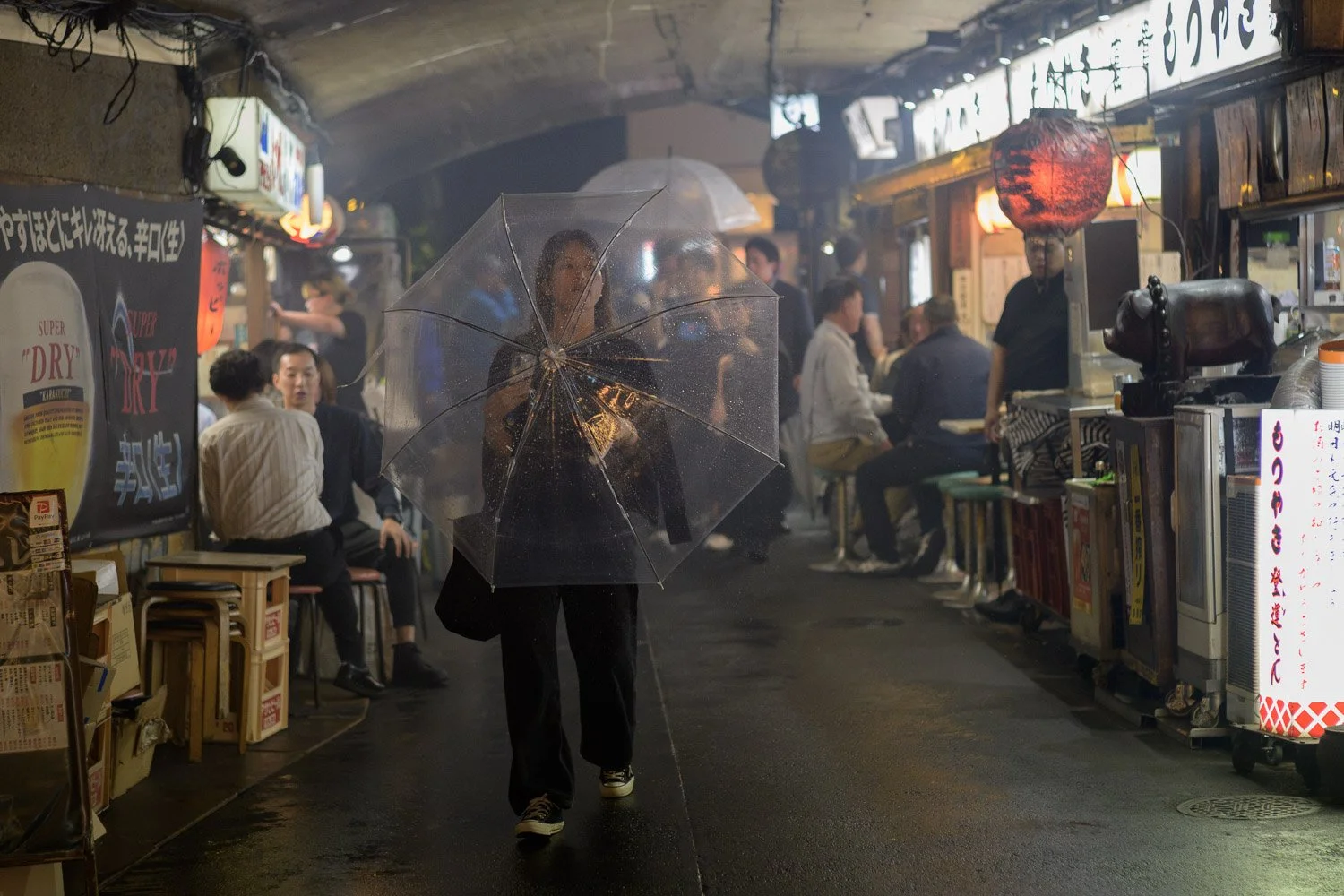 A woman walks through the smoky lantern-lit izakaya lane beneath the railway tracks between Yurakucho and Shimbashi]