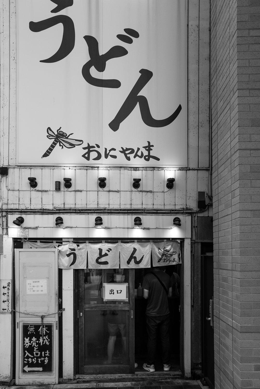 A narrow udon shop front under a huge うどん sign, a customer at the counter inside, Tokyo