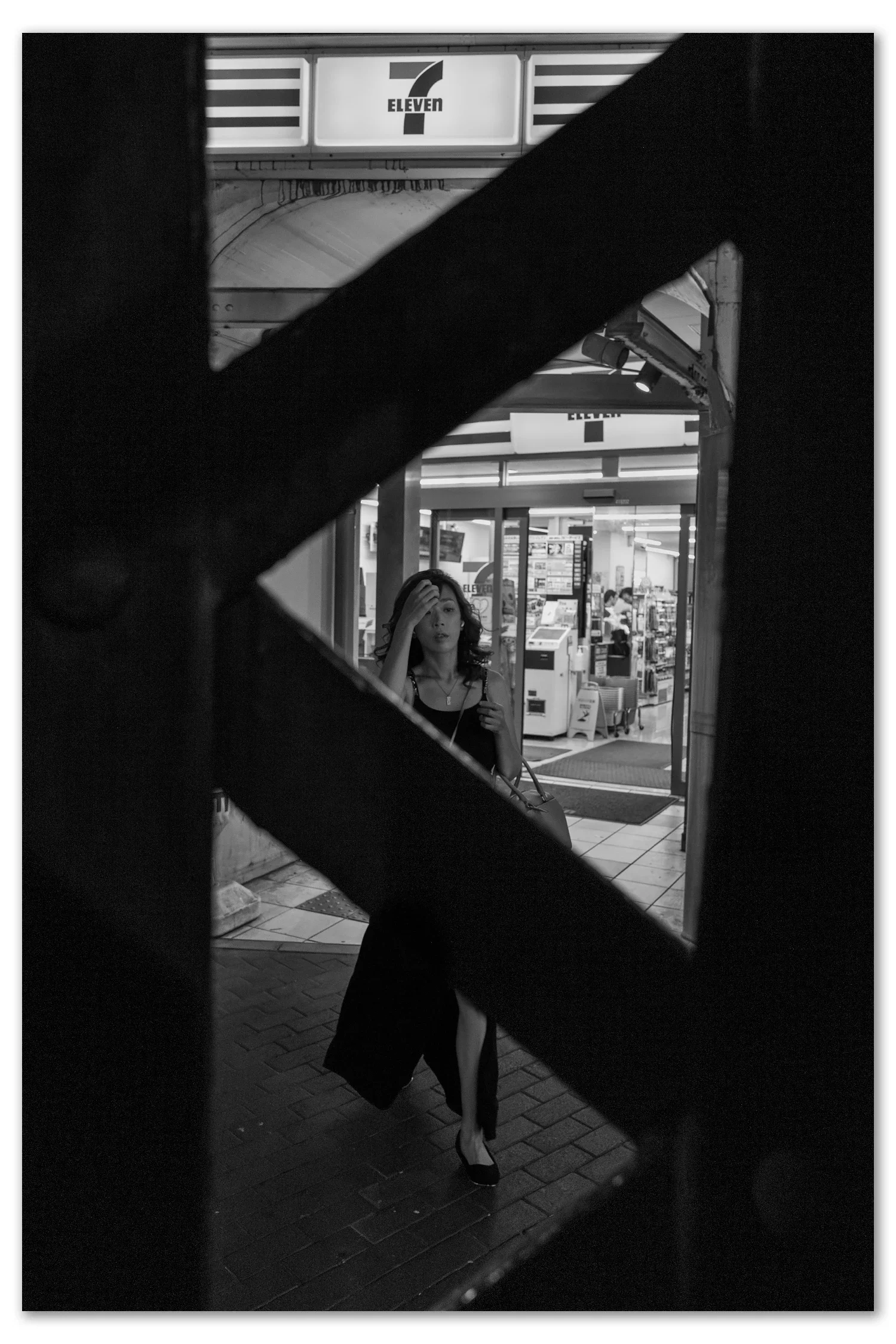 Black and white street photograph, Shinbashi, Tokyo — a woman outside a convenience store framed in the lattice gap of a railway girder, Leica Q2 Monochrom 28mm