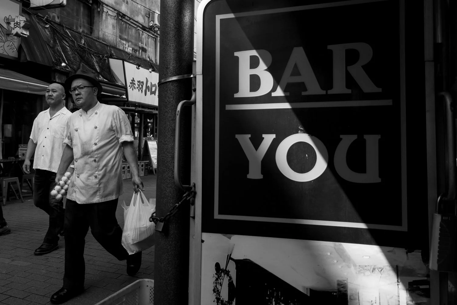 Two cooks in white jackets walk past a BAR YOU sign cut by a slash of hard sunlight, Akabane