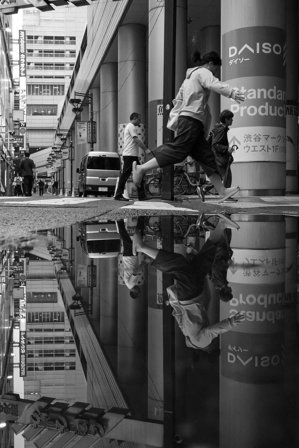 A woman leaps a rain puddle outside Shibuya Mark City, her reflection upside down in the water below, a Daiso sign on the pillar behind her