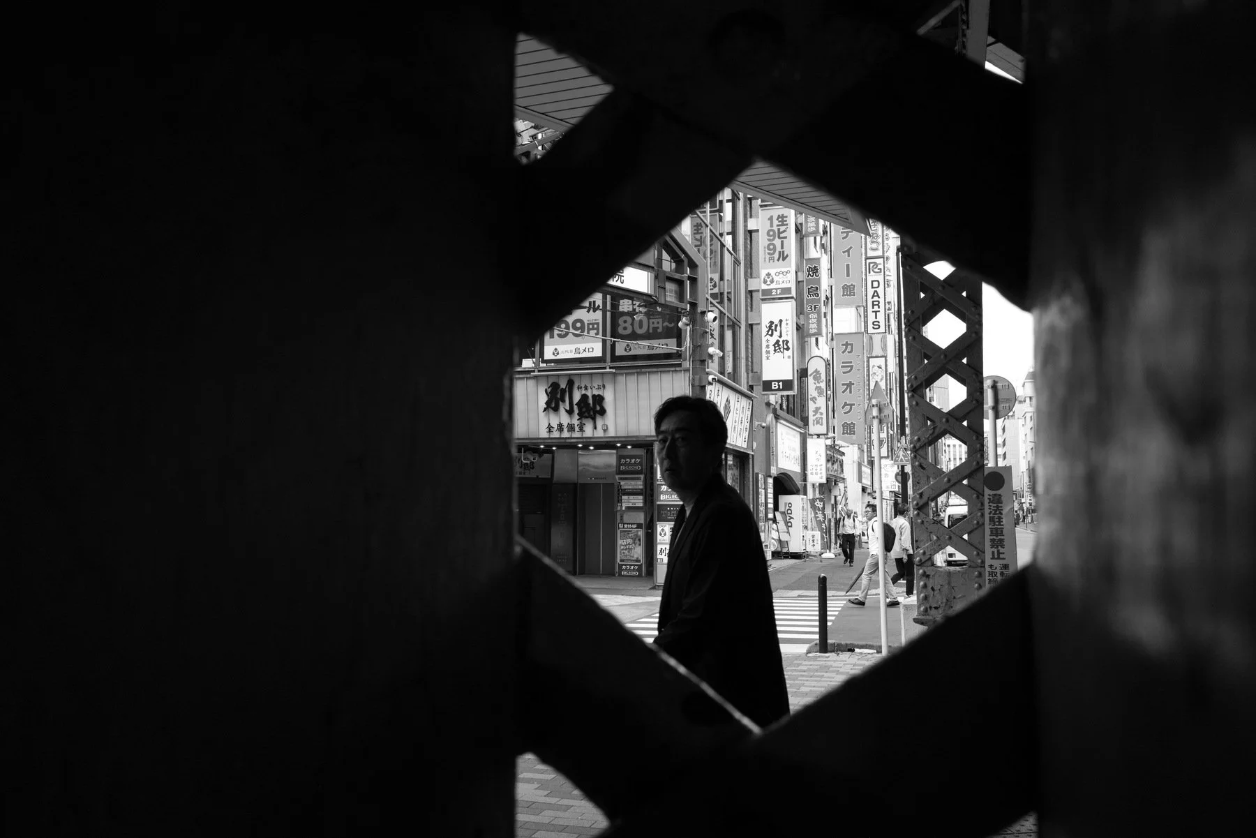 Black and white street photograph, Kanda, Tokyo — a figure silhouetted through an X-shaped gap in a railway girder, Leica Q2 Monochrom 28mm