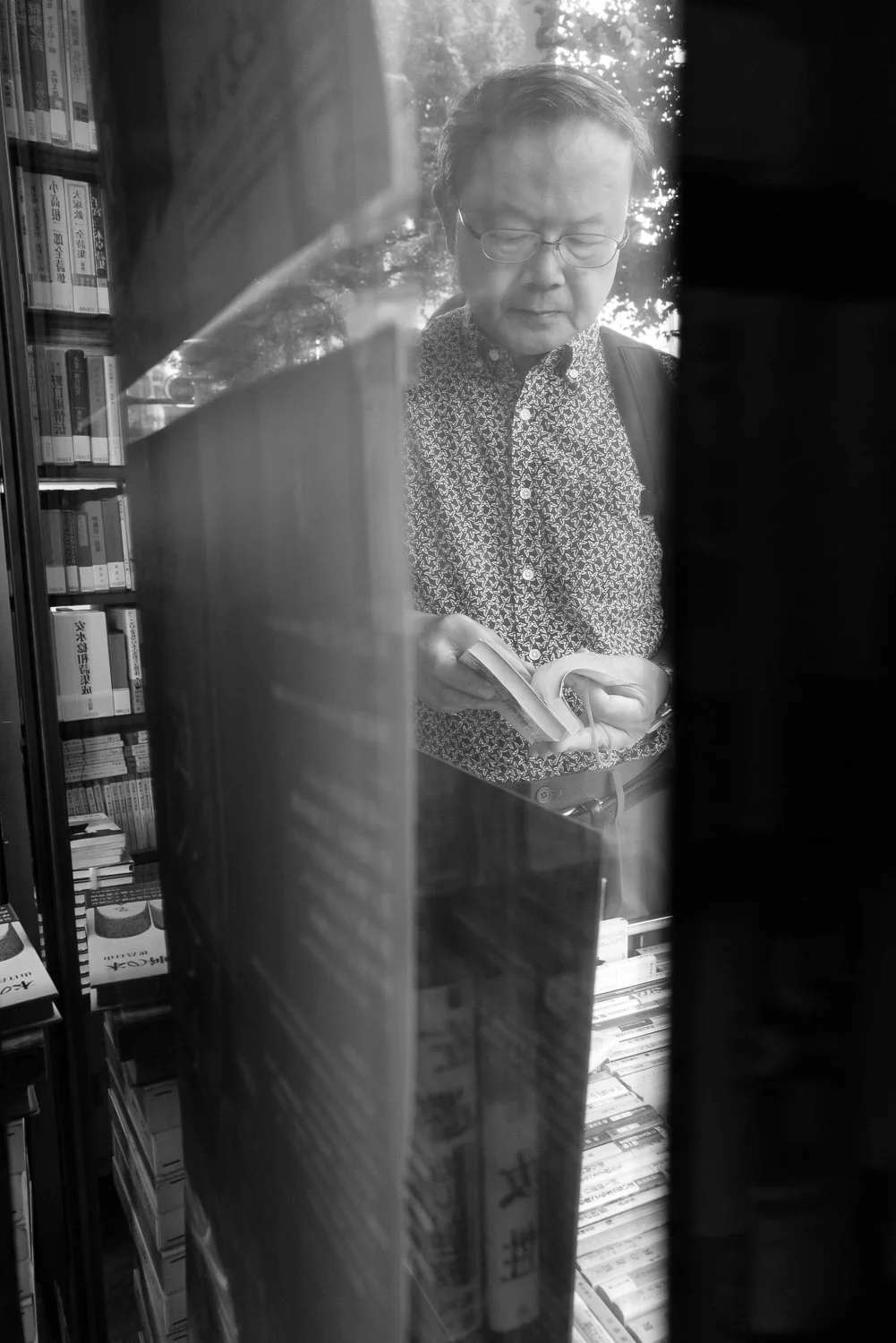 An older man in a patterned shirt reads a paperback between the shelves of a secondhand bookshop, seen through the shop window, Jimbocho