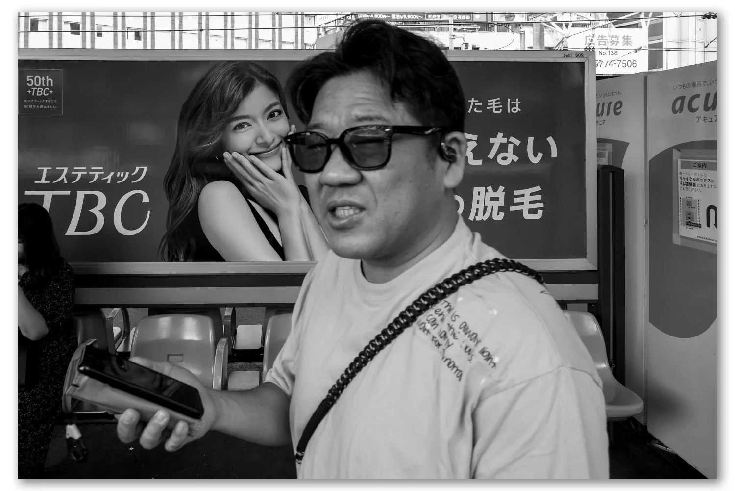 Black and white street photograph, Gotanda, Tokyo — a man in sunglasses passes a beauty-salon billboard at the station, Leica Q2 Monochrom 28mm
