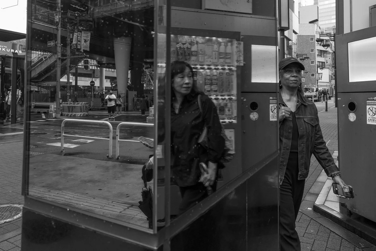 Two women pass a glass shelter beside a vending machine, the Shibuya Sakura Stage sign and a pedestrian deck behind them, Shibuya