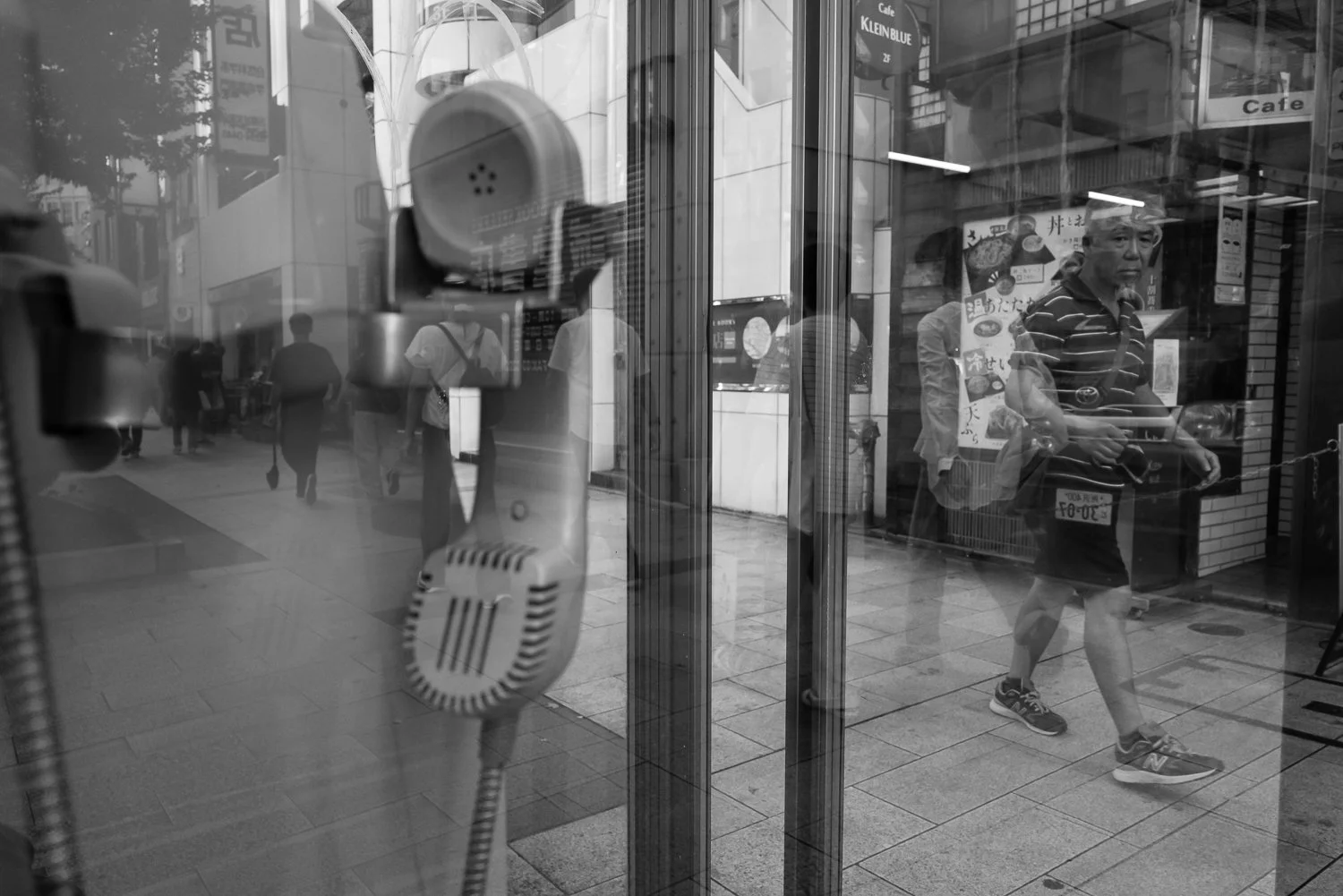 A man passes behind a window reflection of an old telephone handset and a café sign, Tokyo