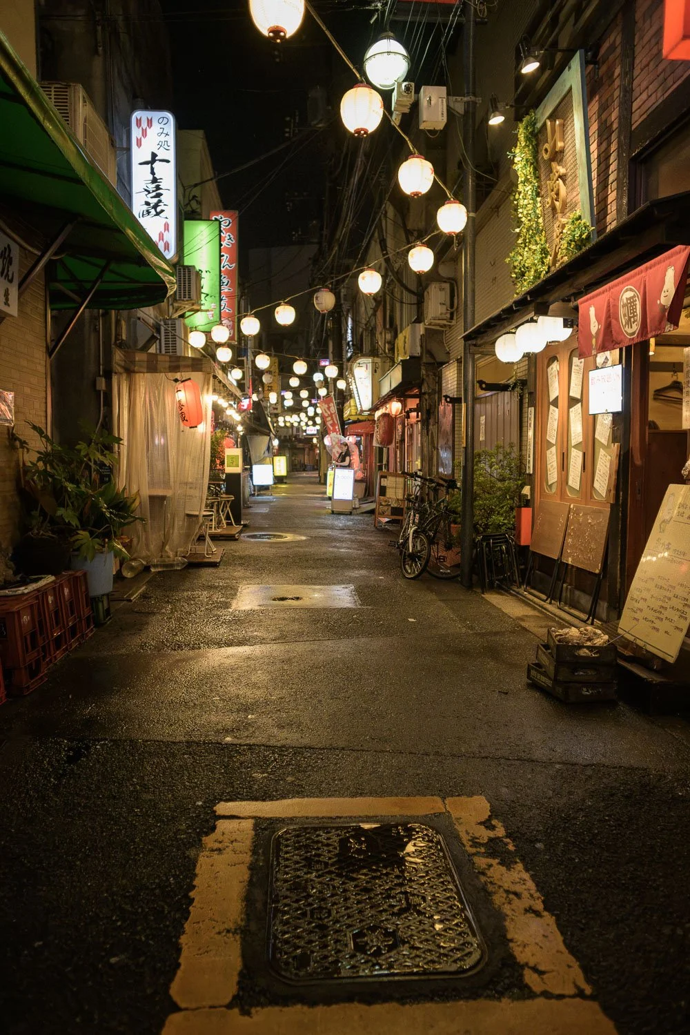 An empty OK Yokocho in Akabane at night, paper lanterns strung above wet pavement and shuttered bars