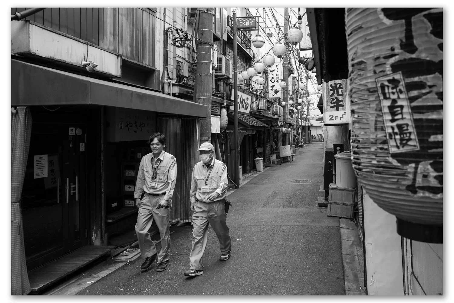 Two older men in short-sleeved shirts walking down a narrow lantern-hung alley in Akabane