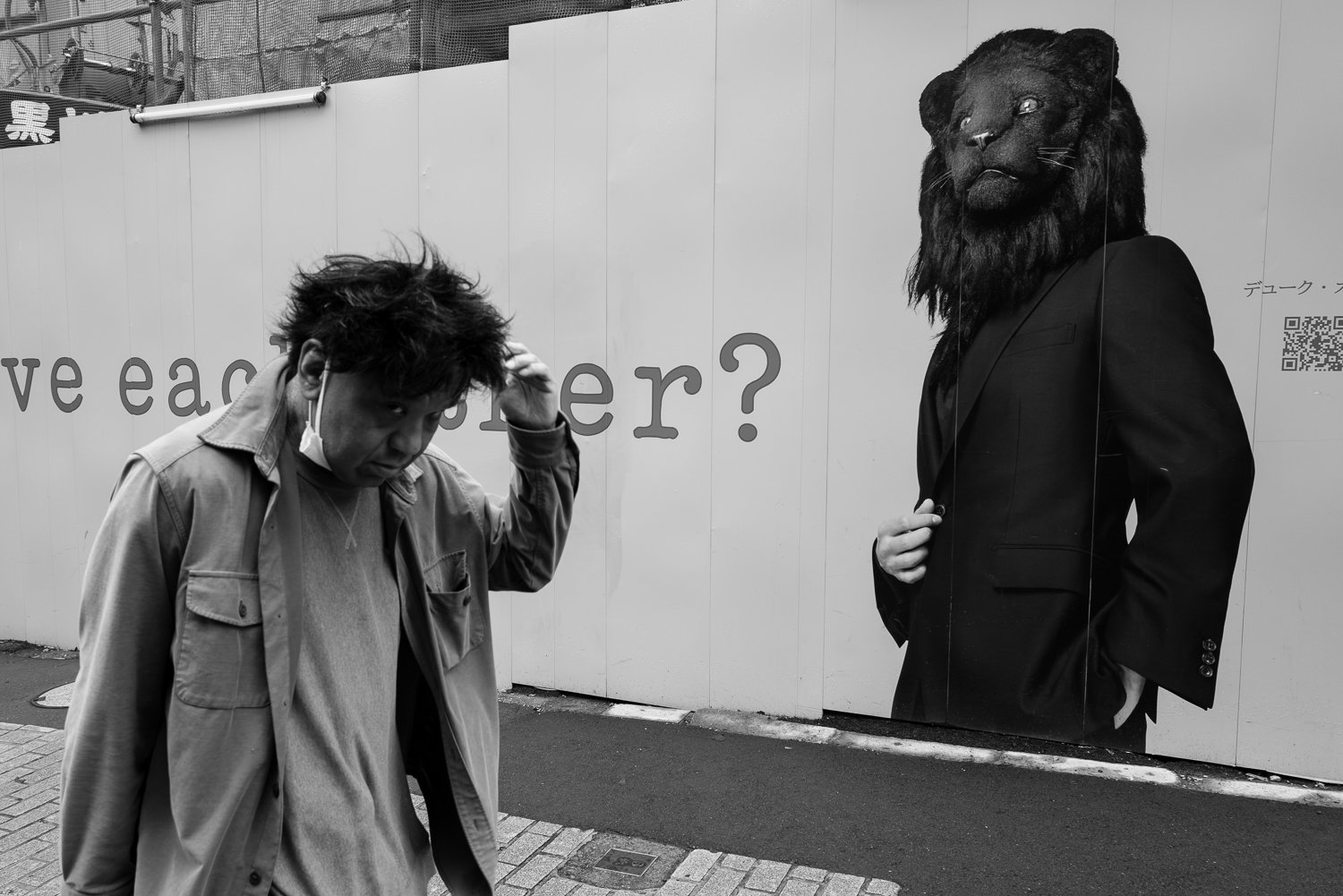 A man pushes his hair back as he walks past a construction hoarding printed with a lion-headed figure in a suit and the words "love each other?", Shibuya